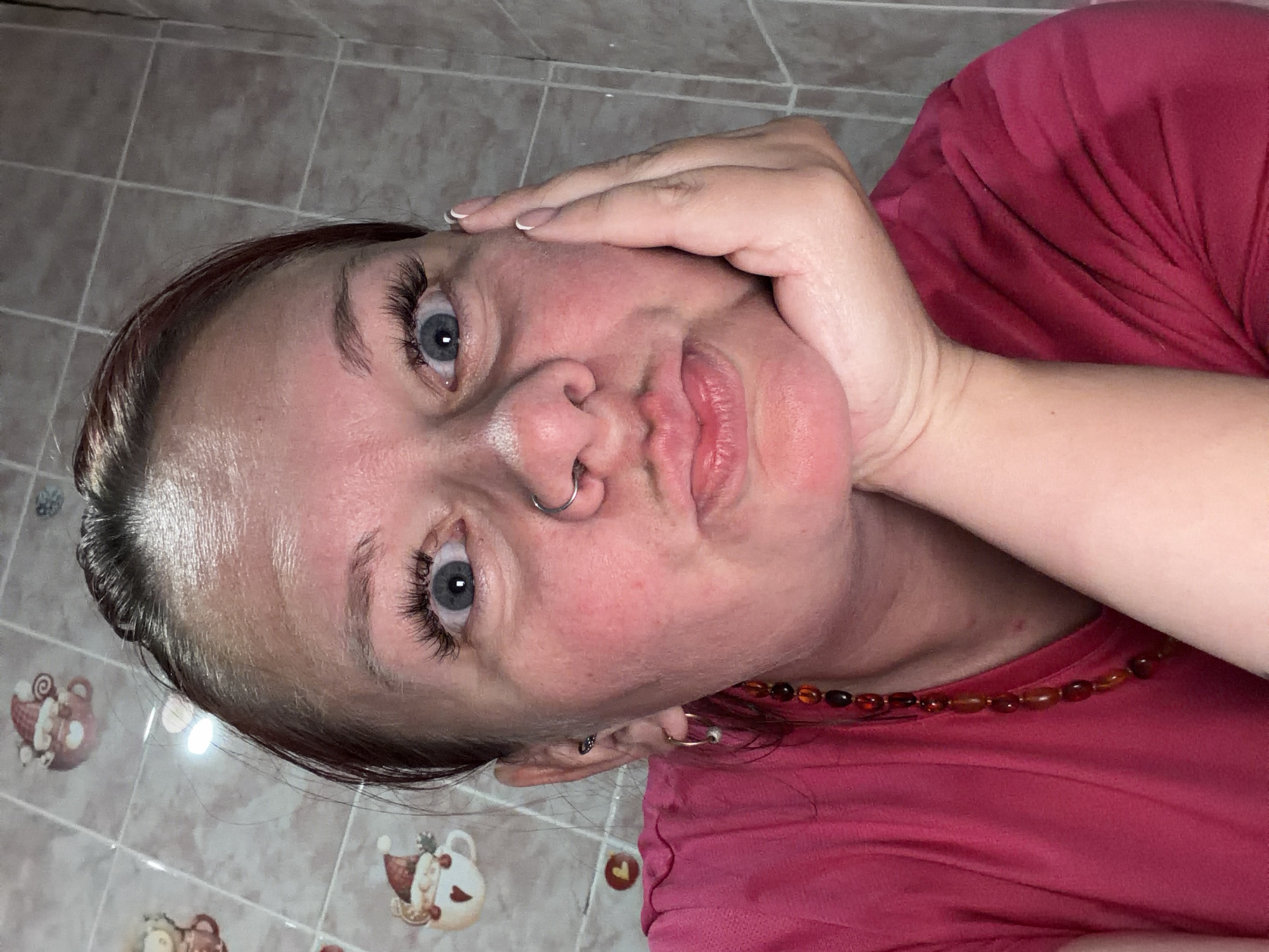 A close-up, rotated portrait features a woman with prominent blue eyes and long, dark eyelashes. Her dark hair is pulled back, and her face shows some redness on the cheeks and forehead. She has a small silver septum piercing and full, slightly pursed lips. Her right hand, adorned with light-colored fingernail polish, is resting against her cheek and jawline. She is wearing a red or maroon t-shirt and a necklace made of amber-colored beads. Several earrings are visible on her left ear. The background consists of light-colored tiled surfaces, possibly a floor or wall, with a grid pattern. Scattered across the tiles are decorative stickers or decals depicting cartoon-like Santa figures and cups, suggesting a festive or cozy indoor setting. The lighting appears to be artificial. There is no visible text.