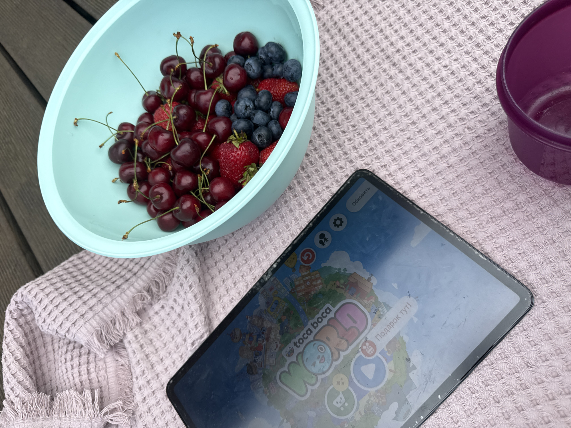 A high-angle shot captures a serene outdoor setting featuring a vibrant bowl of fresh berries, a tablet, and other items arranged on a light purple blanket. The scene is set on dark wooden decking, suggesting a patio or balcony, possibly in Molodezhnyi, Russia.
The primary subject is a light blue plastic bowl, brimming with a colorful assortment of fresh summer fruits. Red cherries, some still attached to their green stems, are abundant, mixed with dark blue blueberries and several ripe red strawberries. The berries appear fresh and appealing.
Next to the fruit bowl lies a black-framed tablet. Its screen displays the "Toca Boca World" game, with the distinctive "toca boca" logo and "WORLD" visible. Text in Russian is present on the screen: "Подарок тут!" (translating to "Gift here!") and "Обновить" (meaning "Update") in the top right corner. The tablet screen exhibits some dust and smudges, hinting at regular use.
These items are placed on a light purple or lavender-colored blanket, characterized by its distinct waffle-weave texture and visible fringed edges. Partially visible on the right side of the frame is a dark purple plastic cup or small bowl.
The natural, soft lighting indicates a clear daytime setting, likely late morning or afternoon. The arrangement suggests a casual leisure activity, perhaps a snack break combined with screen time outdoors. No people are visible, but the objects imply human presence and enjoyment of a relaxed moment. The overall impression is one of simple pleasures and outdoor relaxation.