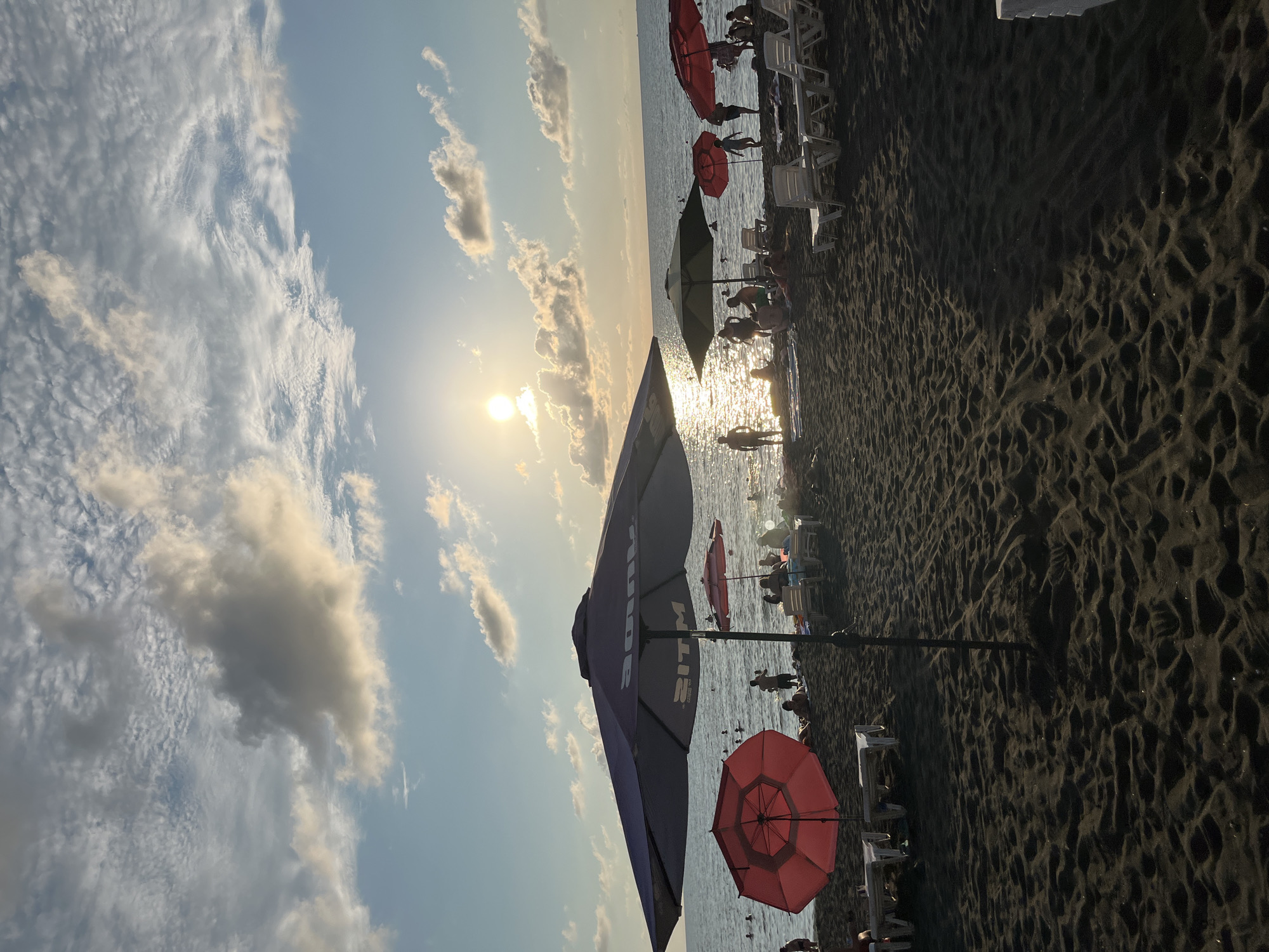 The image depicts a crowded beach in Ureki, Georgia, characterized by its unique black magnetic sand. In the foreground, several beach umbrellas are set up, with a large purple umbrella dominating the left side of the frame. To its left, a vibrant red umbrella stands out. The sand is dark and rippled, showing the texture of the unique magnetic sand Ureki is known for.
In the midground, numerous people are scattered across the beach and in the shallow waters of the sea. Some individuals are standing, while others appear to be relaxing on beach chairs, several of which are visible and are white and empty. People are also seen wading or swimming in the sea. The sea itself is calm, reflecting the bright, late afternoon or early evening sun, creating a shimmering effect on the water's surface.
The sky above is a mix of blue and white clouds, with the sun low on the horizon, casting a warm glow and long shadows across the sand. The overall impression is one of a popular, bustling beach on a pleasant day, taking advantage of the natural beauty and unique sand of Ureki.