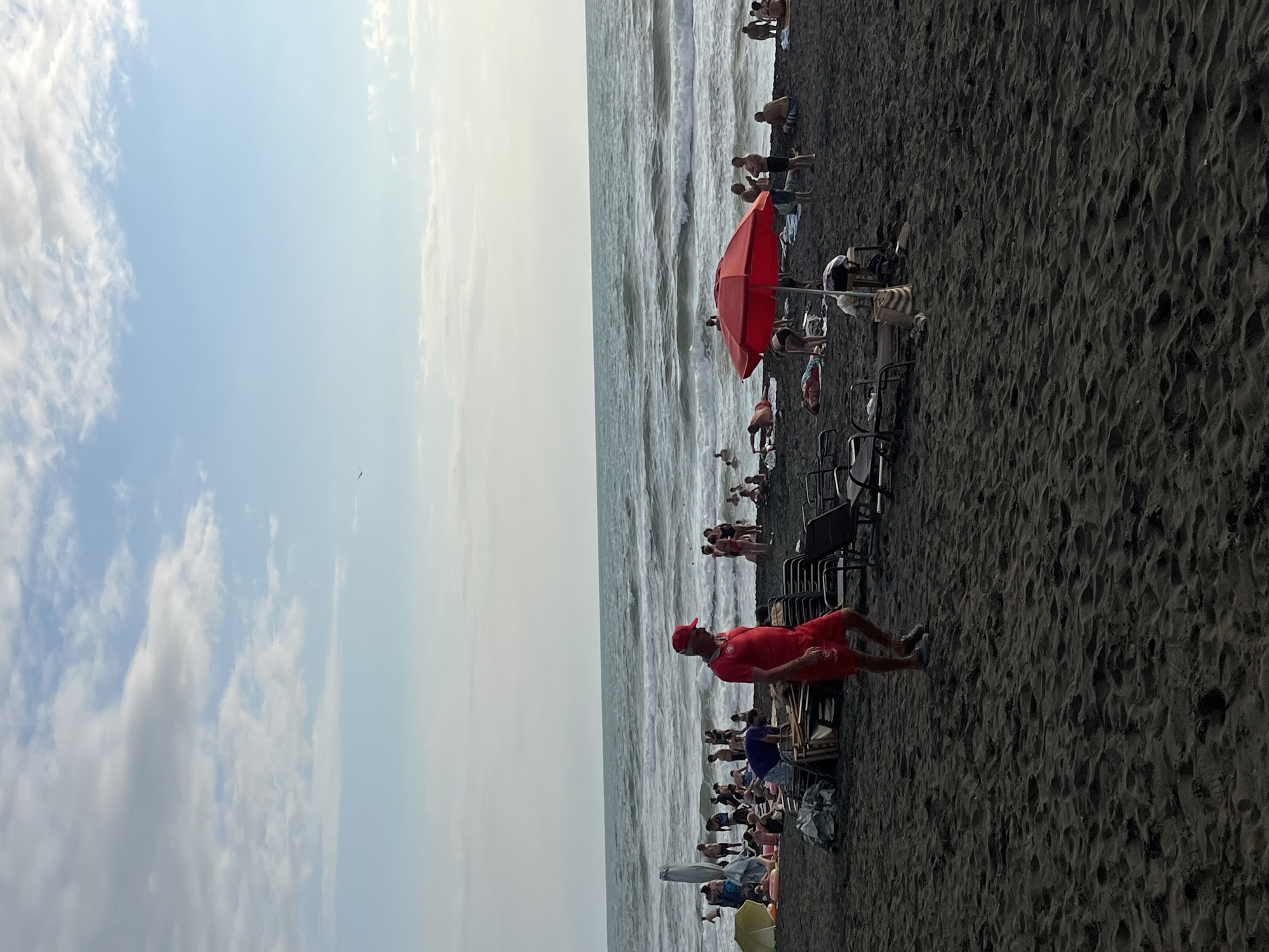 The image depicts a beach scene in Ureki, Georgia, characterized by dark volcanic sand and a calm sea. In the foreground, a man in a red shirt and shorts, possibly a lifeguard, walks on the sand. Several beach chairs and a red umbrella are scattered on the beach. A group of people are visible in the background, some swimming in the sea, while others are relaxing on the sand. The sky is partly cloudy, with a few birds flying in the distance. The overall atmosphere is one of a typical summer day at the beach.