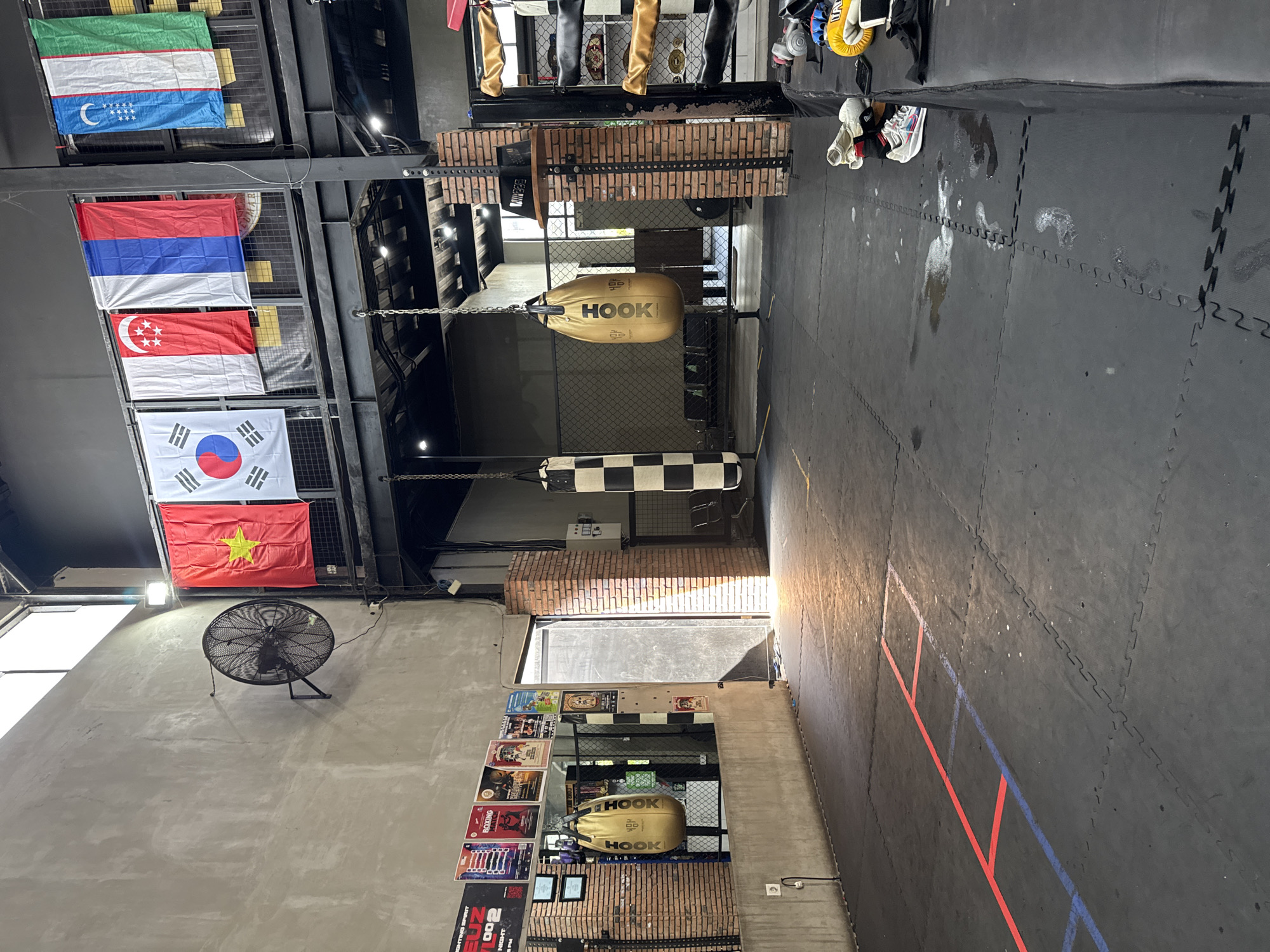 The image depicts the interior of a combat sports gym, likely in Mengwi, Indonesia based on the flags displayed. The gym features black rubber mat flooring with faded red and blue painted markings. Several punching bags hang from the ceiling, including a tan "HOOK" branded bag, and a black and white checkered bag. Flags representing Uzbekistan, Serbia, Singapore, South Korea, and Vietnam are displayed on the wall. A large fan is mounted on a gray concrete wall adjacent to a display of posters. The gym appears to be well-used with visible scuff marks and stains on the floor. Natural light enters from an open doorway. The overall environment suggests a space for training and fitness.