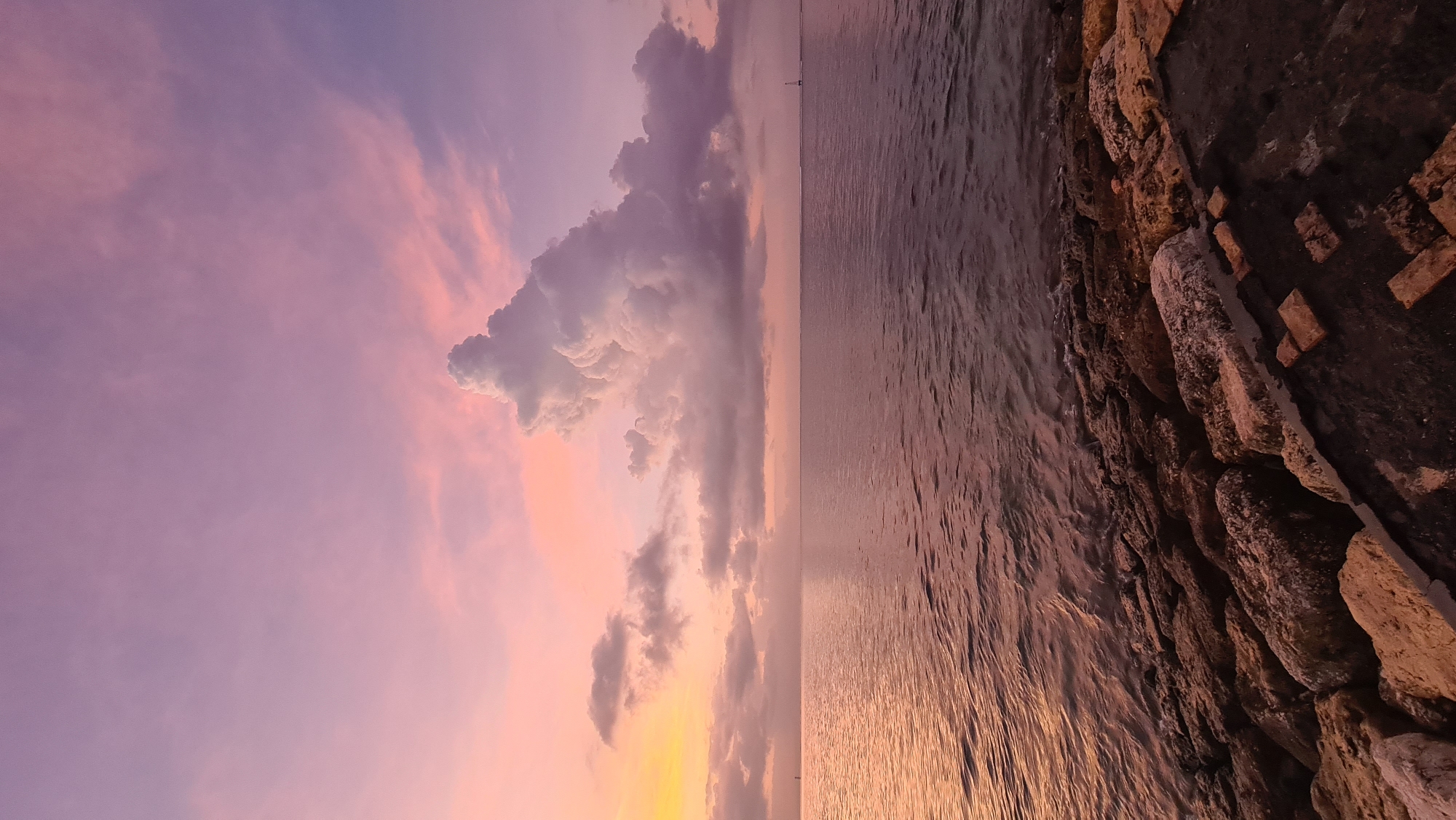 The image depicts a sunset scene by the sea in Denpasar, Indonesia. The sky is awash in soft hues of pink and purple, with fluffy clouds catching the last rays of the sun. The ocean stretches out to the horizon, its surface reflecting the colors of the sky, creating a serene and peaceful atmosphere. On the right side of the frame, a stone embankment lines the shore, with rough-hewn rocks and some scattered bricks, suggesting a man-made structure protecting the coastline. The lighting indicates it is either sunrise or sunset, with the warm glow illuminating the scene. There are no people or specific objects prominently featured, making the natural elements of the sky and sea the main subjects. The overall mood is tranquil and contemplative, capturing the beauty of a tropical evening.