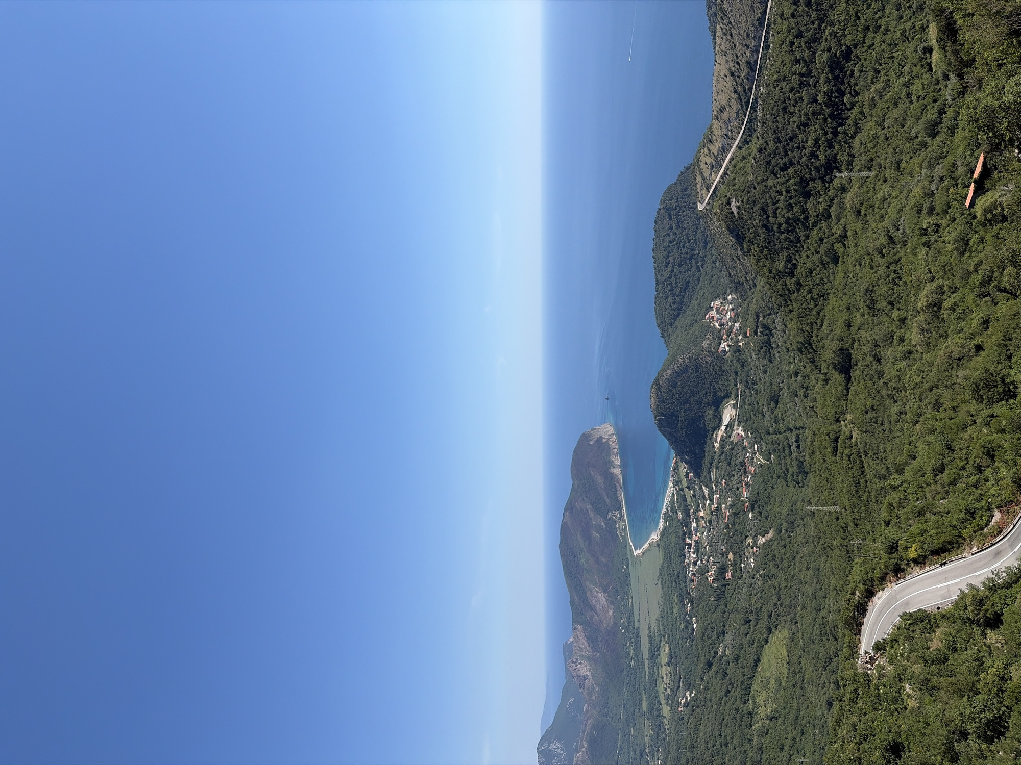 The image presents a panoramic view from a high vantage point overlooking a coastal region. In the foreground, a winding paved road cuts through lush green, densely forested hills. The road features white dividing lines and appears to be in good condition. Further into the middle ground, the landscape transitions to a flatter area with scattered buildings, forming a small village. Beyond the village, a crescent-shaped sandy beach lines the azure blue sea. The sea stretches to the horizon, meeting a clear, bright blue sky with no clouds visible. In the distance, mountainous terrain frames the coastline. The scene appears to be devoid of people. This view is consistent with overlooks found in the vicinity of Novoselje, Montenegro, a location known for its scenic coastal and mountainous landscapes.
