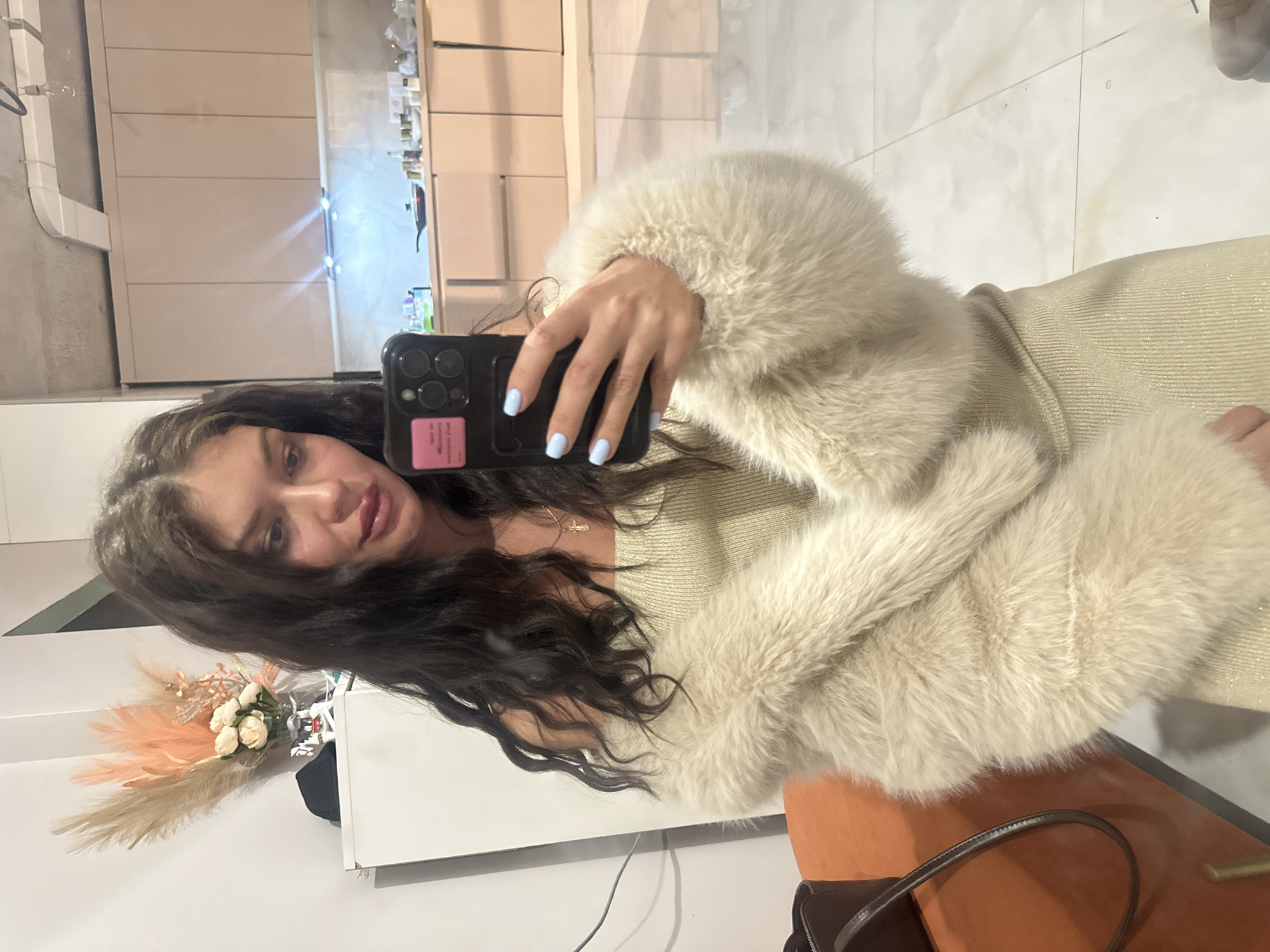 A young woman is depicted in a mirror selfie, looking towards the camera with a subtle smile. She has long, dark, wavy hair and is wearing a light-colored, possibly beige or champagne-toned, shimmery top or dress. Over this, she sports a fluffy, cream or off-white faux fur coat with voluminous sleeves. Her fingernails are painted a light blue. She holds a dark-colored smartphone, appearing to be an iPhone with multiple cameras, in a matching dark case that features a small, unreadable pink sticker on its back.
The indoor setting reflected in the mirror reveals light-colored tiled flooring and sleek, light beige wall paneling or cabinetry in the background. Bright reflections from overhead light sources are visible on the surfaces. To the left side of the reflection, decorative elements include a bouquet of pampas grass in shades of beige and peach, alongside white faux flowers, placed on a surface. On the right, a section of an orange or brown upholstered chair or bench, and a dark handbag with a brown strap are visible. The image is presented rotated 90 degrees clockwise from its natural portrait orientation. The location is Khimki, Russia.