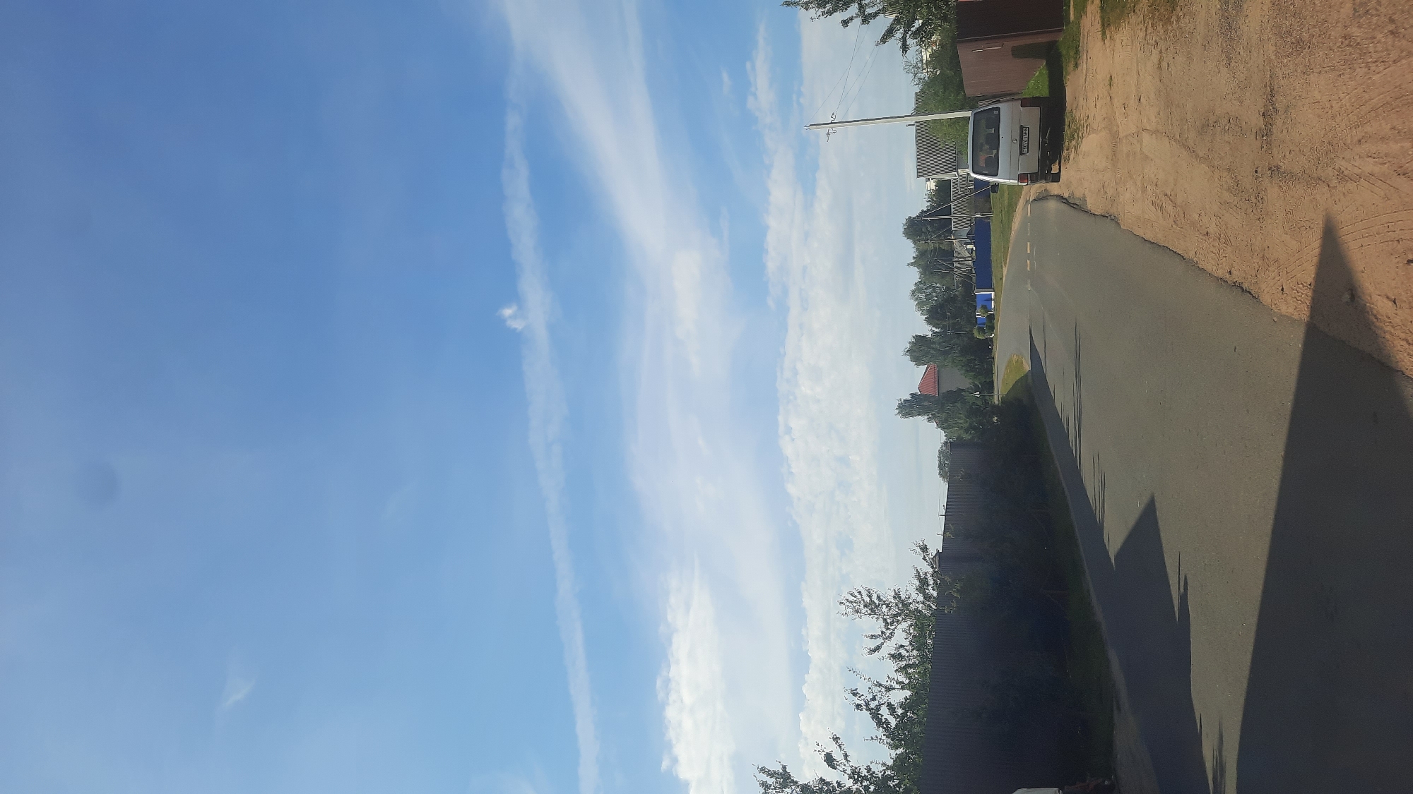 The image depicts a suburban street scene in Shklov, Belarus, under a bright blue sky with scattered white clouds. A paved road curves to the right, bordered by a sandy verge on the right and a dense hedge behind a metal fence on the left. A light-colored van is parked on the sandy verge. Beyond the fence and hedge, several residential buildings with red roofs and grey siding are visible, interspersed with trees. A utility pole stands on the right side of the road. The lighting suggests it is daytime, likely late morning or afternoon, casting shadows from the trees across the road.