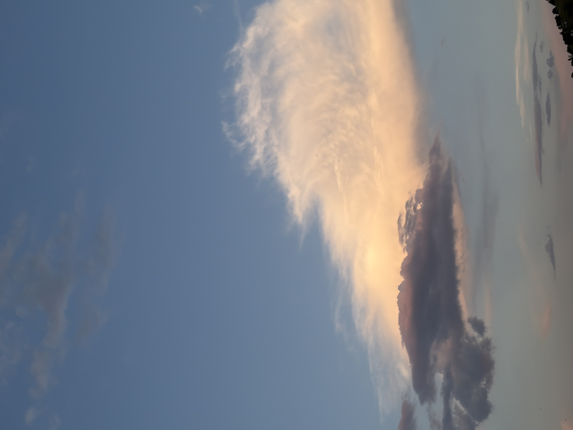 The image shows a sky with clouds. The sky is predominantly blue, with patches of white and gray clouds. Some of the clouds have a pinkish hue, possibly due to the setting sun. The image appears to have been taken at dusk or dawn. The location is unspecified.