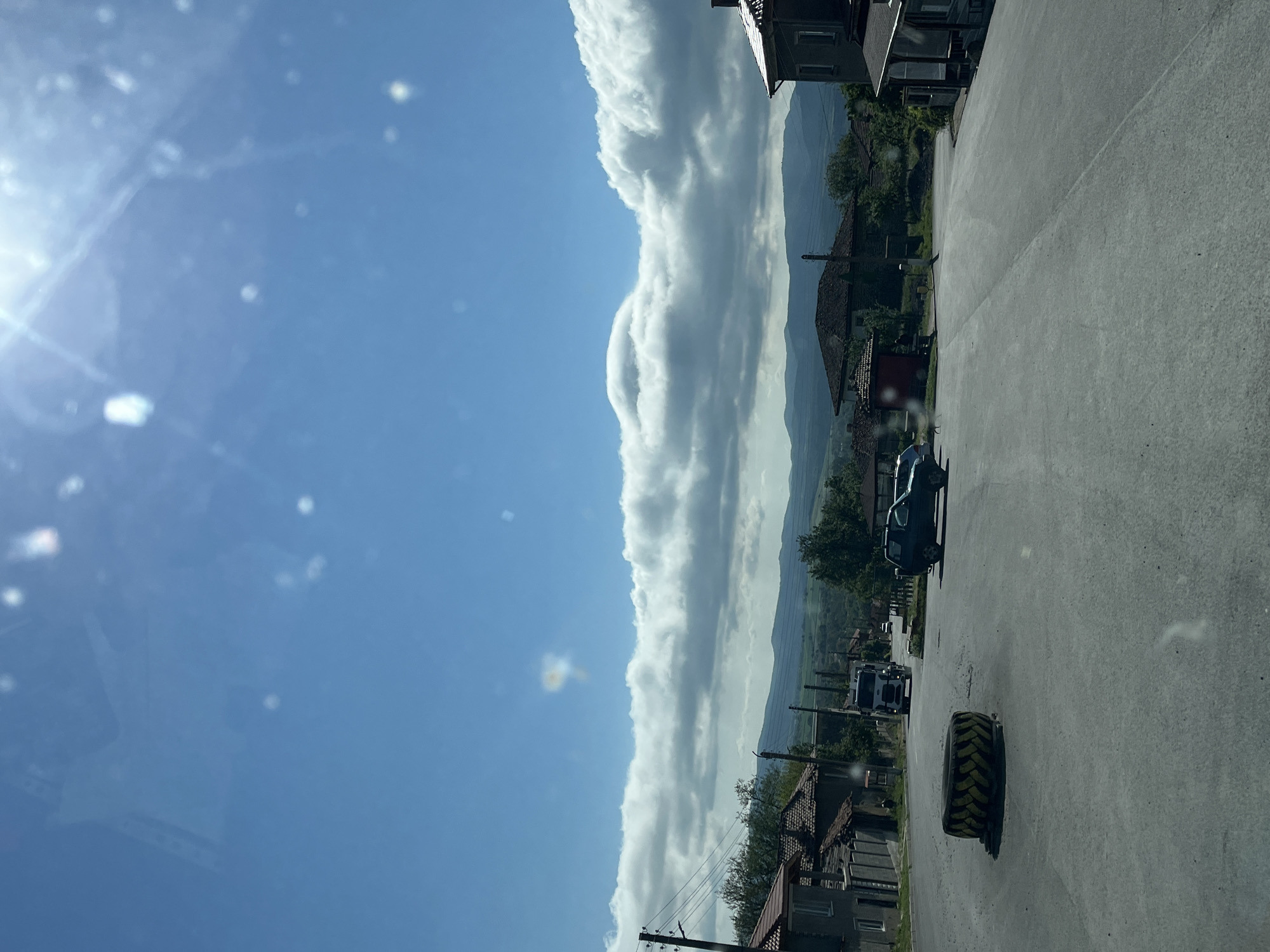 The image depicts a street scene in Rish, Bulgaria. The sky is predominantly blue with scattered clouds. A line of puffy, elongated clouds stretches horizontally across the midground. The foreground is a gray asphalt road, with a large tractor tire lying on the side. Several vehicles are parked along the side of the road, including a dark-colored car and a white truck. Buildings are visible on either side of the road, with red tile roofs and gray facades. Mountains can be seen in the distance behind the buildings. Power lines are also visible. The overall perspective suggests the image was taken from within a vehicle.