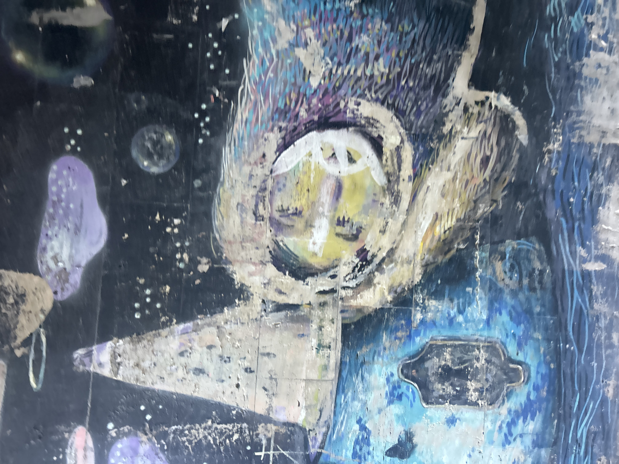 This is a close-up, high-angle, full-shot, color photograph of a painting on a rough surface, possibly a wall or a canvas. The painting depicts a stylized face with large, closed eyes, rendered in yellow and white, set against a dark, textured background. The face appears to be partially obscured by flowing, colorful brushstrokes in shades of blue, purple, and white, resembling hair or headdress.
To the left of the face, there are abstract, bulbous shapes in purple and pink, dotted with white specks that could represent stars or glitter. A light-colored, pointed object, perhaps a beak or a wing, extends from the lower left towards the center of the image. To the right of the face, the painting transitions into vibrant blues and dark hues, with a metallic, dark-colored object embedded within the blue area, resembling a plaque or a decorative element. The overall style is abstract and somewhat surreal, with visible brushwork and signs of wear or texture on the surface. The lighting is even, with no discernible time of day or weather cues. There is no visible text in the image.