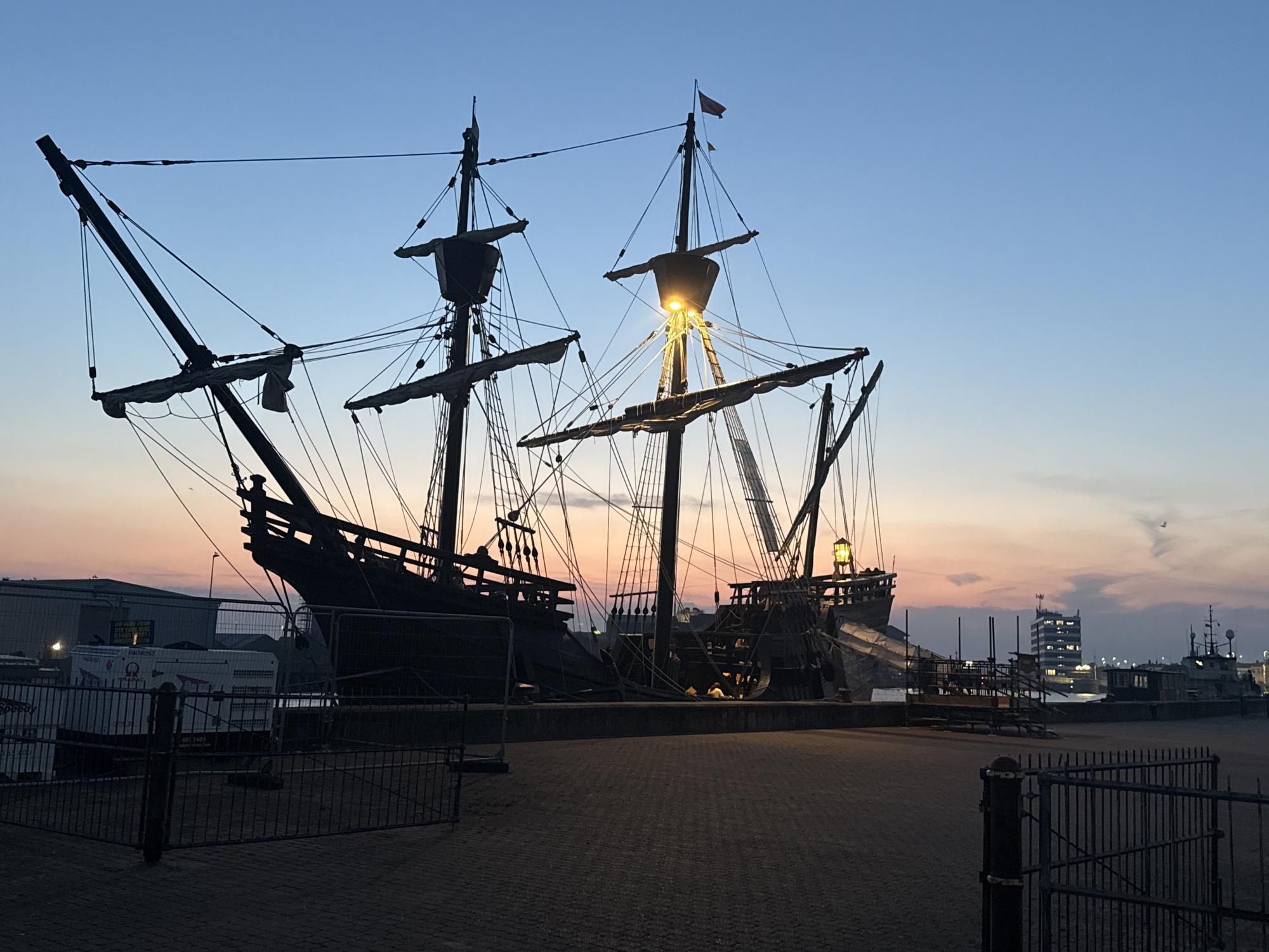 This is a medium shot, taken at dusk, of a replica of the 17th-century ship the 'Golden Hinde' docked in Great Yarmouth, United Kingdom. The ship is silhouetted against a gradient sky, transitioning from a pale blue at the top to hues of orange and pink near the horizon. The ship itself is dark and imposing, with its three masts and rigging clearly defined. Several lights are visible on the ship, including a bright yellow light illuminating the crow's nest on the main mast. The ship is positioned on a paved area, with metal fencing in the foreground. In the background, several other vessels are docked, and a modern office building with illuminated windows stands further inland. The overall scene is peaceful, with the setting sun casting a warm glow over the maritime setting.