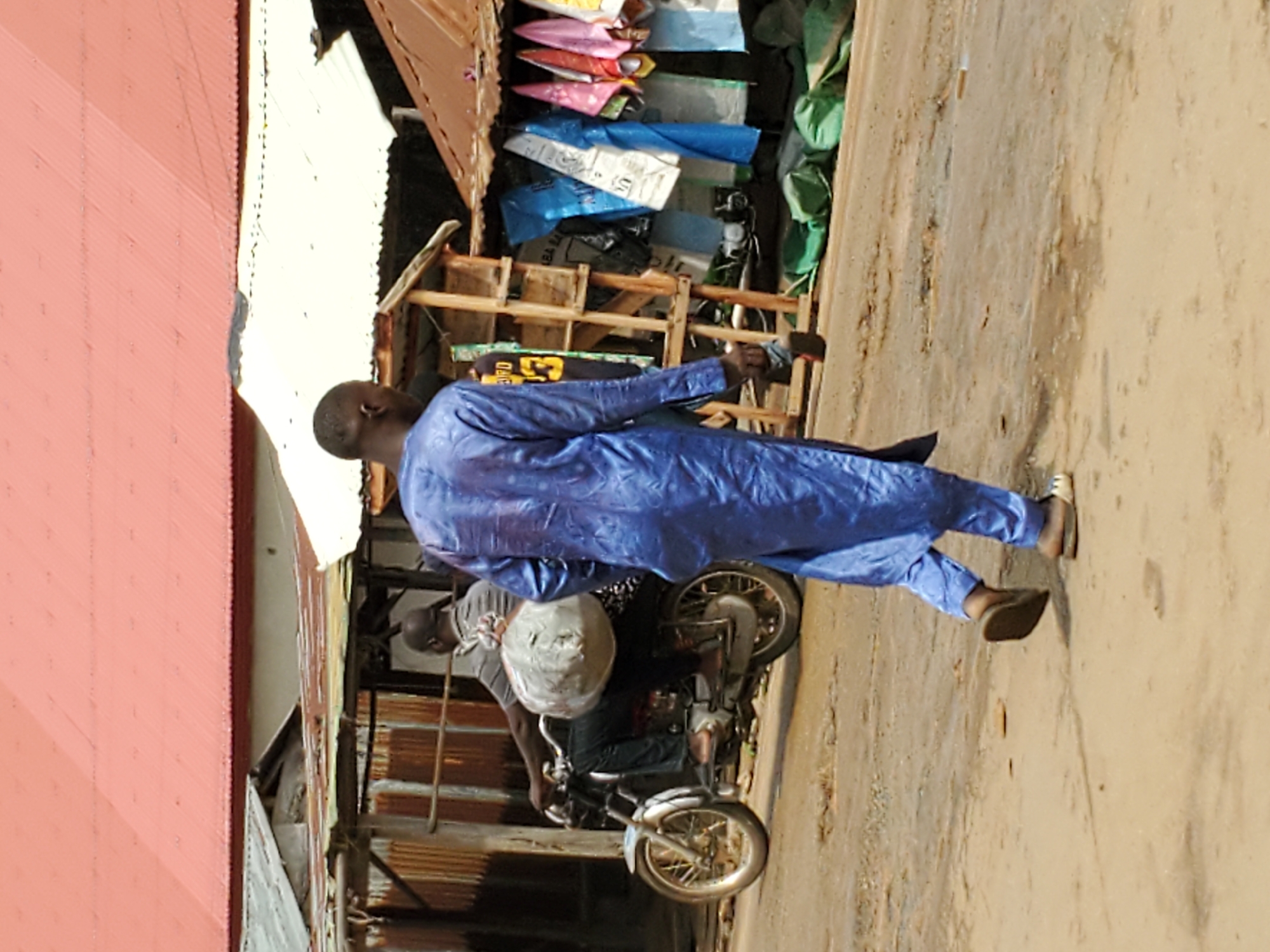 The image, oriented vertically, depicts an outdoor street scene in Mararaba, Nigeria, captured during daylight hours. The central figure is a man walking away from the viewer towards the right side of the frame. He wears a long, flowing blue traditional garment with a subtle patterned texture, and light-colored sandals on his feet. His head is closely shaven or has very short dark hair.
Slightly behind and to the left of the walking man, another person is positioned on a motorcycle. This individual is seen wearing a grey t-shirt and dark trousers, with a white helmet or similar object resting on the motorcycle. The motorcycle features spoked wheels.
On the right side of the scene, behind the walking man, there is a market stall or small shop constructed from wood and corrugated metal. Its shelves display various goods, notably items packaged in brightly colored plastic bags, including pink, blue, and green.
To the far left of the image, a building with a reddish-orange corrugated metal roof is visible. The ground is an unpaved, light brown dirt path. The bright, clear lighting indicates a sunny day. No clearly legible text is discernible within the image. The setting suggests a lively commercial or residential street.