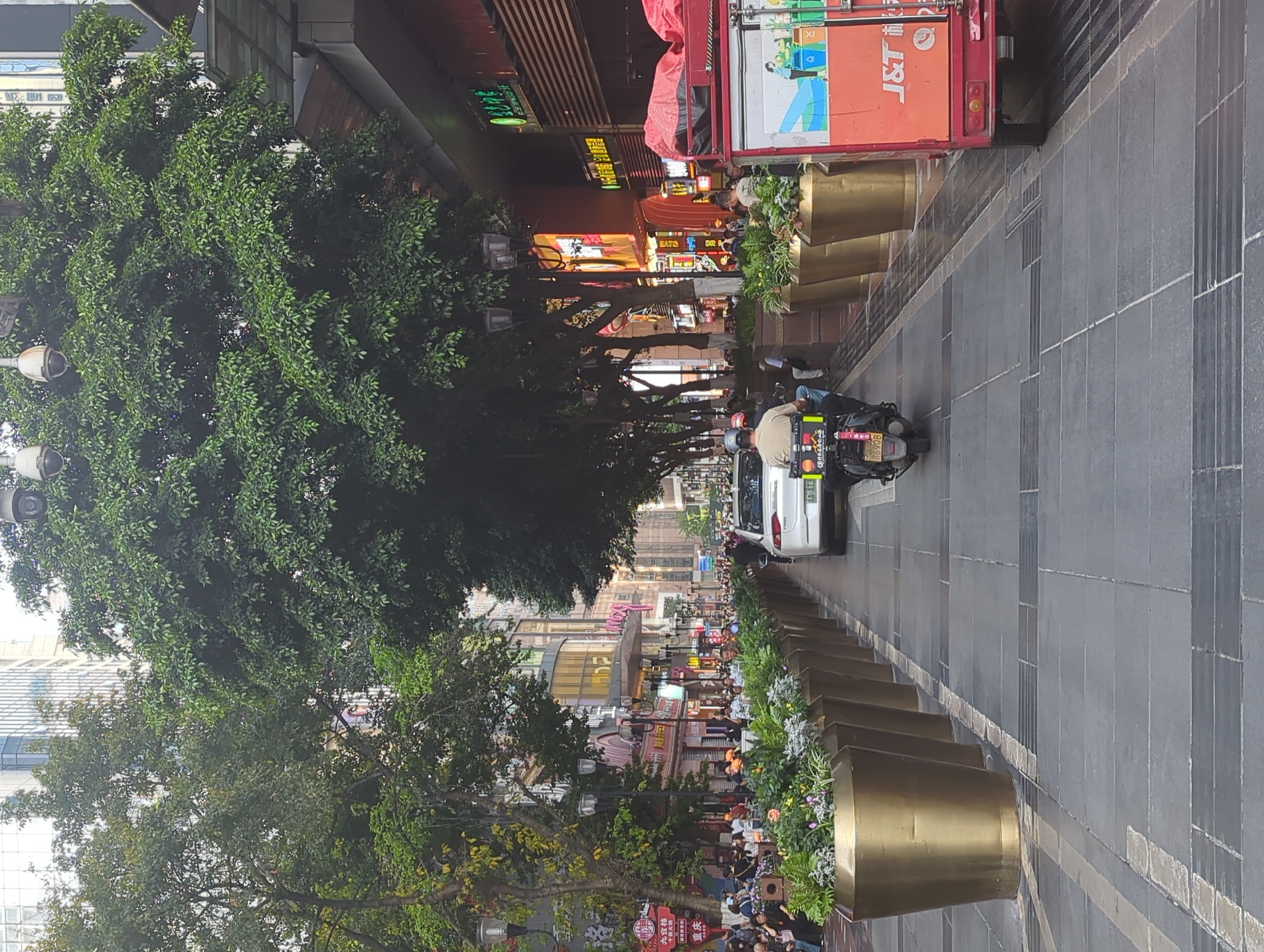 The image depicts a bustling urban street scene in Yuzhong District, China, likely a commercial or pedestrian area. The street extends into the distance, flanked by multi-story buildings and a dense line of mature green trees on the left.
Numerous people are visible, walking and gathering along the paved area and near storefronts. In the mid-ground, two individuals are riding a white scooter or motorcycle on what appears to be a mixed-use roadway. Behind them, part of a white car is visible. On the right side of the street, a red J&T Express delivery truck is parked, facing away from the viewer.
The street is well-maintained with dark grey paving stones and lighter grey strips. A series of large, golden cylindrical planters filled with lush greenery and flowers lines the right side of the paved area, acting as a divider. Streetlights are integrated into the tree line.
The buildings feature various commercial facades, some with large glass windows and others with illuminated signs, suggesting active businesses. The overall atmosphere appears lively and typical of a busy city street during daytime, possibly late afternoon, given the ambient light and some illuminated signs.
Visible text includes "J&T Express" in English and Chinese on the delivery truck. The scooter has a license plate "86908" and some Chinese characters. Several shop signs display Chinese text, such as "正宗" (authentic) and partially visible "EATS". A red banner in the foreground on the left reads "九优街" (Jiǔ Yōu Jiē - Nine Excellence Street) and "重庆" (Chóngqìng - Chongqing).