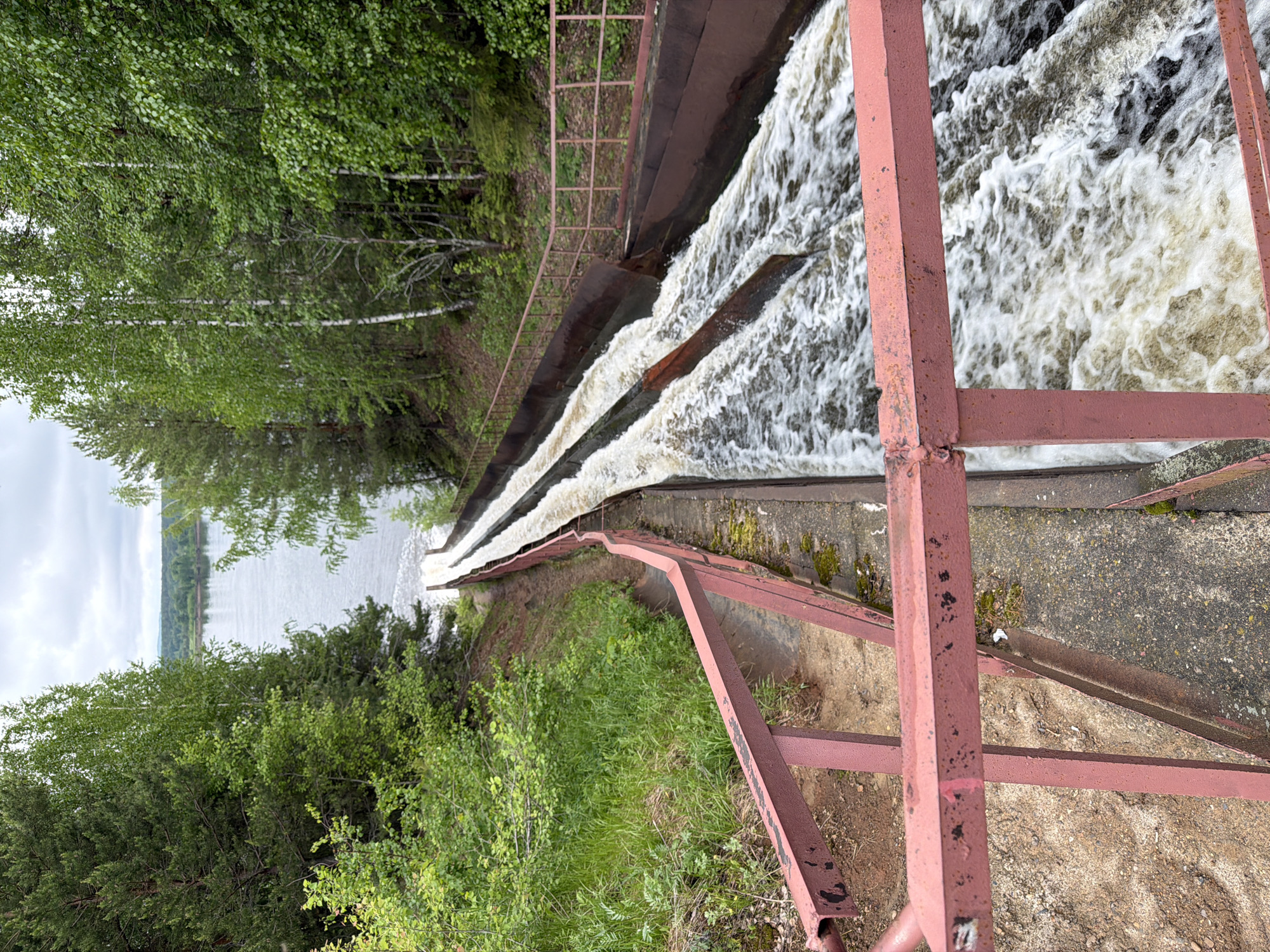 View from an elevated position in Chernushka, Russia, overlooking a man-made waterfall structure. Water cascades over a concrete dam, creating turbulent white water. A rusted reddish-brown metal safety railing runs alongside the dam. Lush green trees border the left side of the image, contrasting with the concrete and metal structures. In the distance, a body of water and a hazy horizon are visible. The weather appears overcast. The composition emphasizes the dam and waterfall, suggesting an observation point.