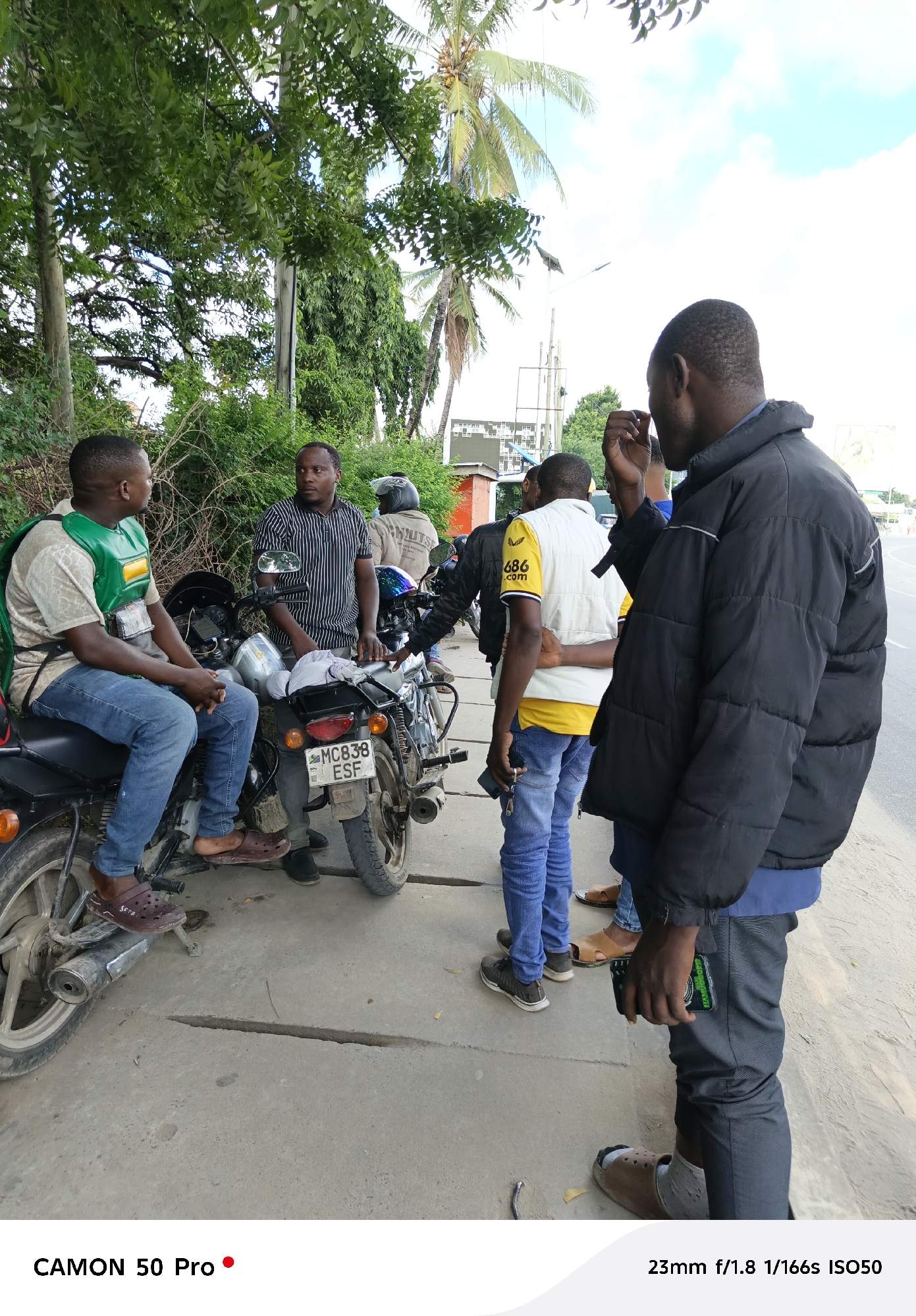 The image captures a casual gathering of several African men on a sidewalk next to a road. Two individuals are seated on motorcycles, while others stand around them engaged in conversation. The license plate of one of the motorcycles is visible and reads "MC838 ESF". Clothing worn by the individuals varies, including jeans, t-shirts, jackets, and casual footwear like sandals and crocs.
In the background, lush green vegetation and palm trees are visible. A building can be seen with what looks like posters and a power pole with transformers. The scene appears to be in an urban environment. Based on the license plate and general setting, the location is likely Dar es Salaam, Tanzania.