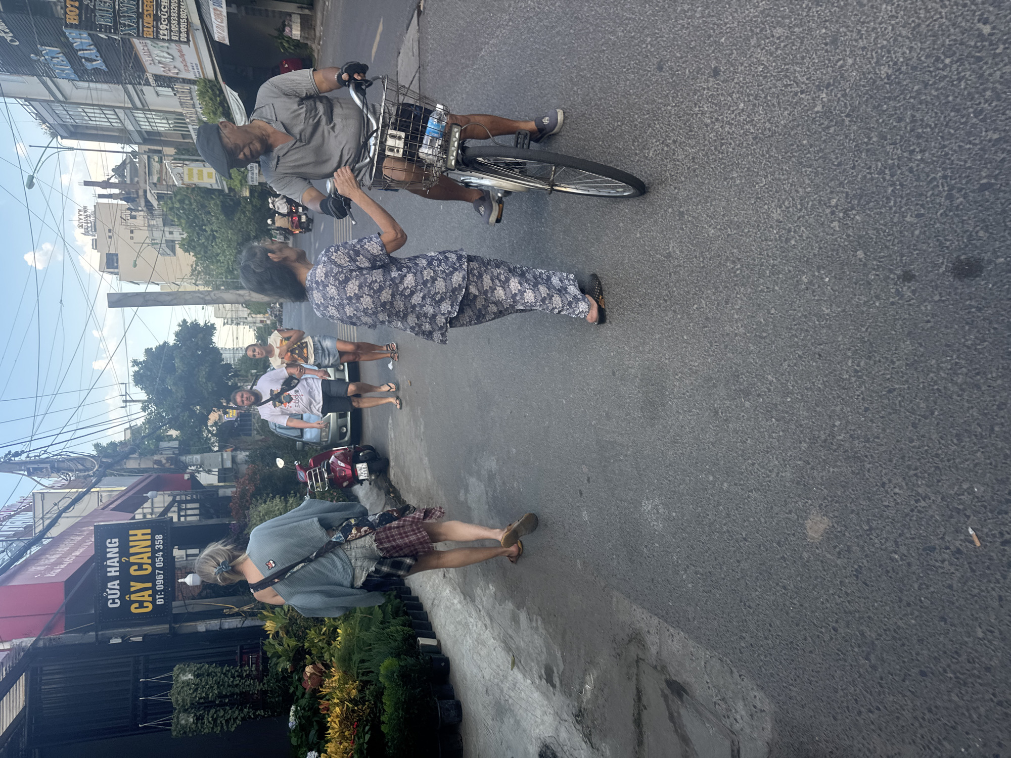 In Bac Nha Trang Ward, Vietnam, on a street lined with shops and trees, a man wearing a grey striped polo shirt and a flat cap rides a bicycle. He is in the process of interacting with an older woman who is wearing a floral patterned shirt and pants and is standing to his left. The woman has her right hand extended, appearing to be gesturing or reaching towards the man. Behind them, another woman, dressed in a blue off-the-shoulder top and patched denim shorts, walks away from the camera. To her left, a red scooter is parked, and further down the street, a man in a white t-shirt and shorts walks away from the camera. The environment includes a sidewalk with potted plants and a paved street. The sky is visible above, with some clouds.