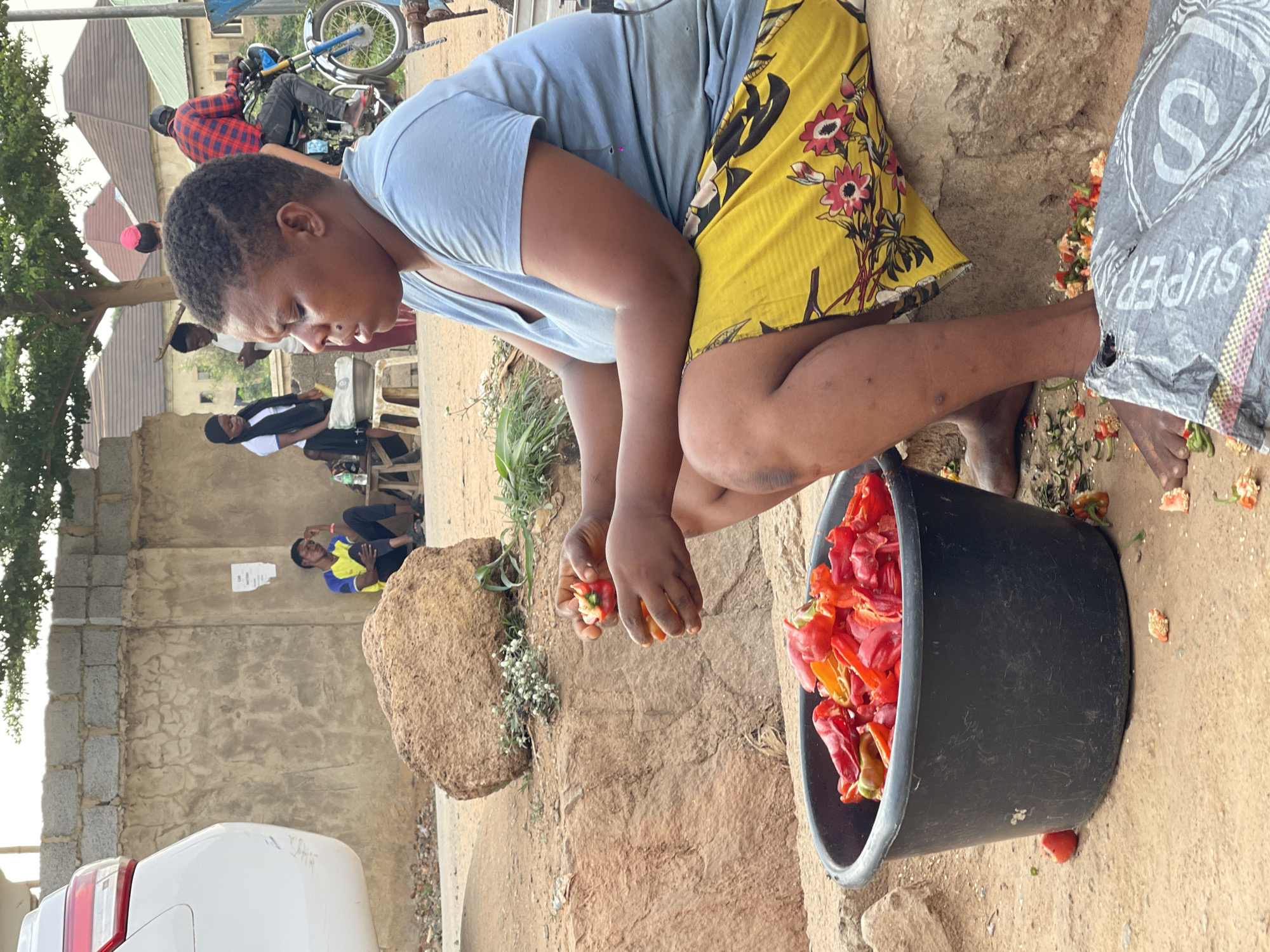 A person squats outdoors near a bucket filled with red and orange peppers. The individual wears a light blue shirt and a yellow skirt with floral patterns. They are holding peppers in their hands, possibly preparing them. The scene is in an urban setting with buildings, vehicles, and other people visible in the background. The location is Abuja, Nigeria. Other objects in view include a rock, a white car, and a "Super" brand bag. People in the background are either sitting or riding a motorcycle. The environment appears to be a mix of dirt, cement, and brick surfaces.