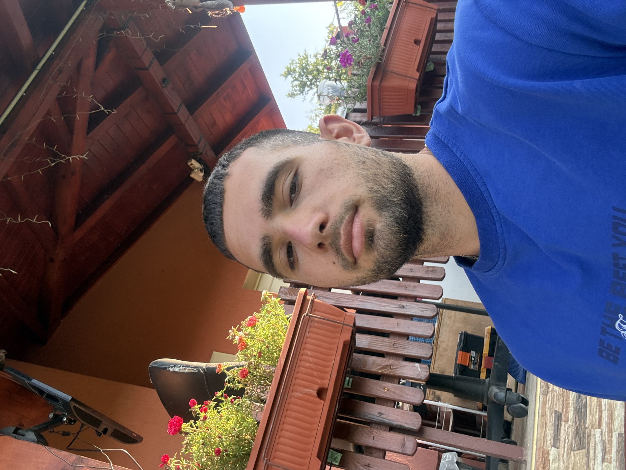 A young man with short dark hair and a beard is taking a selfie outdoors. He is wearing a blue sweatshirt. Behind him, there is a wooden railing and flower boxes with red and pink flowers. The background also shows a building with an orange wall and a wooden roof structure. There is a television mounted on the wall. The scene appears to be in Balinovtsi, Bulgaria.