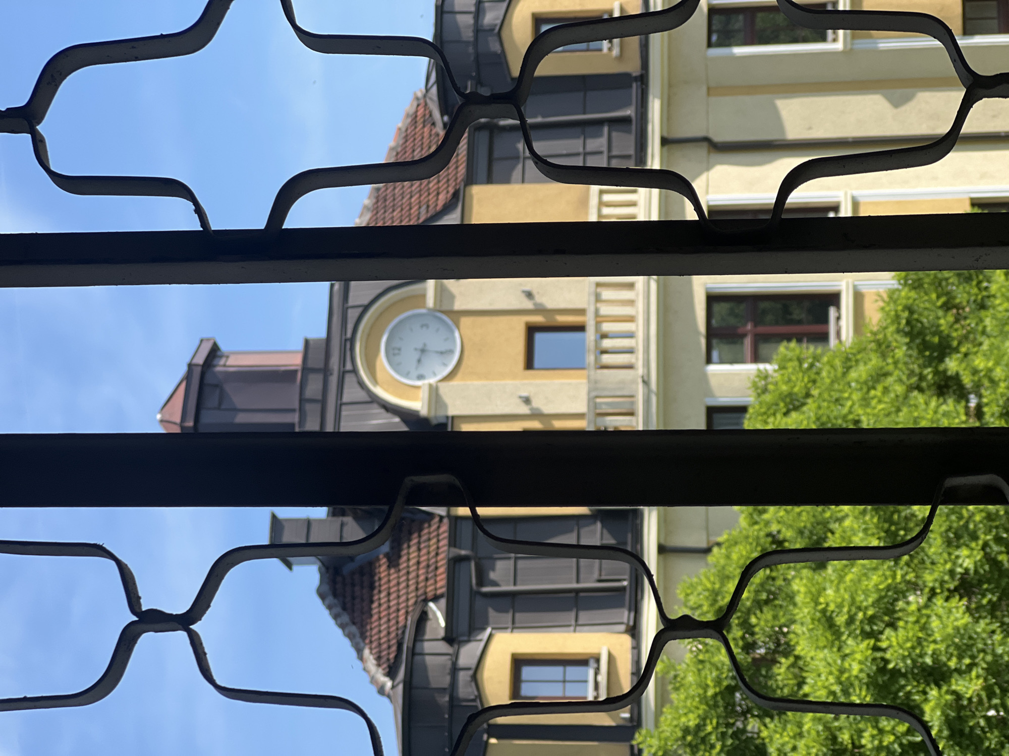 The image presents a view through a metal security grate, revealing an exterior building with a prominent clock tower. The building is painted in shades of yellow and features a combination of architectural details, including a red tile roof, dark metal accents, and rectangular windows. The clock face is white with black numerals and hands. To the right, there is green foliage indicating trees. The sky is visible in the upper left of the frame, indicating the time of day is during daylight hours. The location is in the city of Unknown, Unknown.