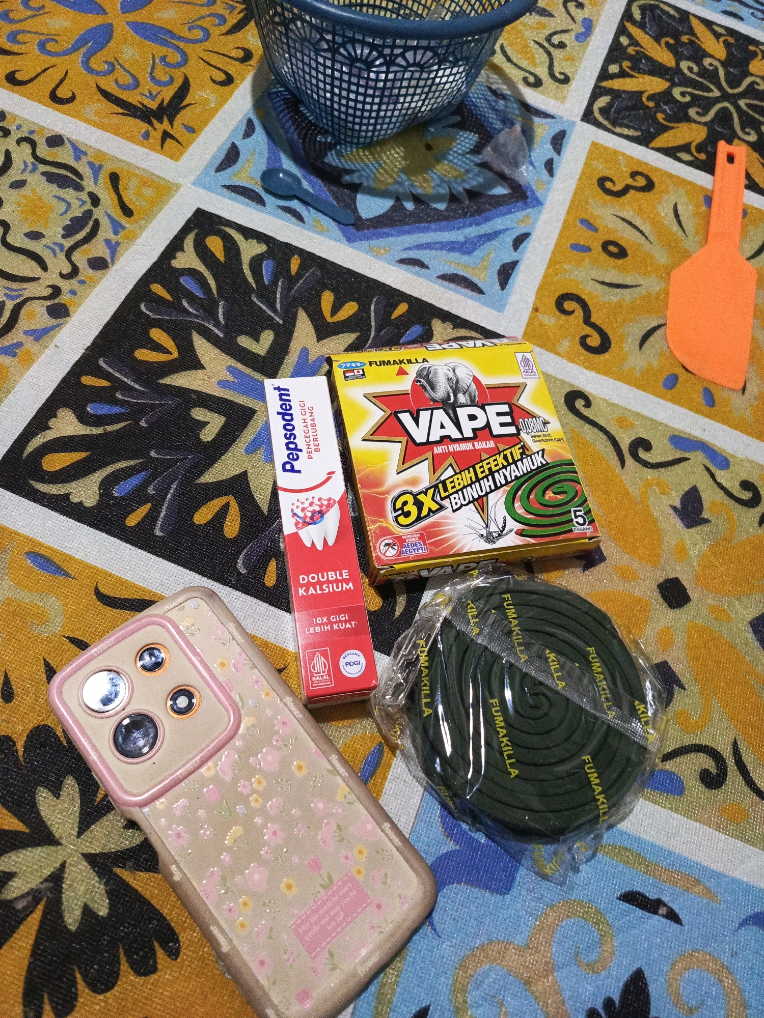 A still life arrangement of household items is laid out on a patterned tablecloth. The tablecloth features a repeating mosaic of yellow, blue, and black floral and abstract motifs.
Prominently displayed are a tube of Pepsodent toothpaste with "Double Kalsium" branding, a box of Vape mosquito coils with an elephant graphic and text in Indonesian, and a sealed package of green mosquito coils. A pink floral patterned smartphone case with multiple camera lenses is positioned in the lower left corner. An orange spatula is visible on the right side of the image. In the upper portion, a blue wire basket is partially visible.
The scene appears to be an indoor setting, likely a table, with no discernible geographical landmarks of Megang Sakti I, Indonesia. The items are static, suggesting a staged photograph rather than an active event.