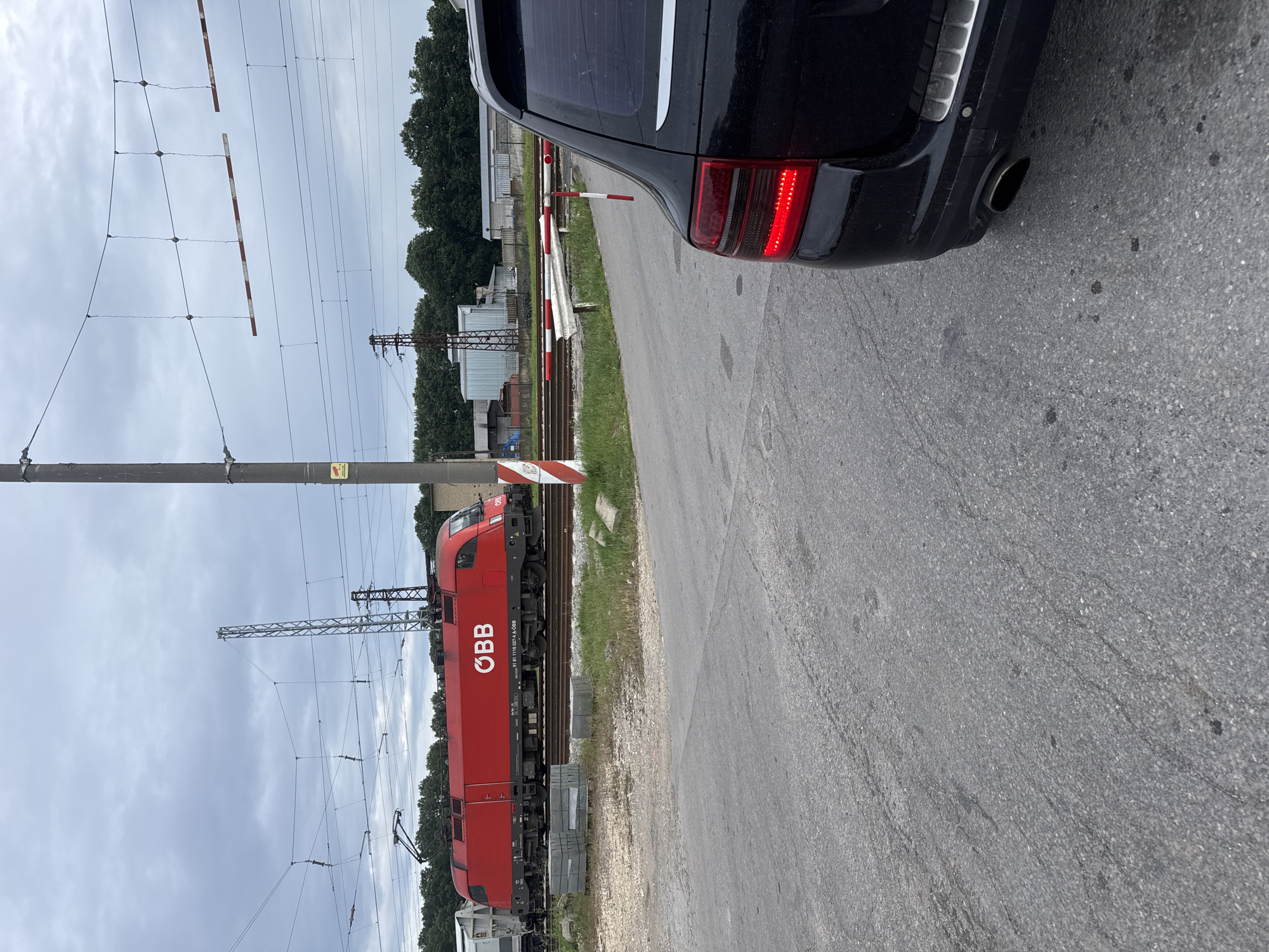 The image shows a red ÖBB (Austrian Federal Railways) electric locomotive at a railroad crossing in Senovo, Bulgaria. The crossing gates are down, indicating the train is passing or about to pass. A black car is stopped close to the crossing, its rear partially visible. The sky is overcast. Railroad tracks, overhead power lines, support poles, and trees are visible in the background. The road surface is asphalt.