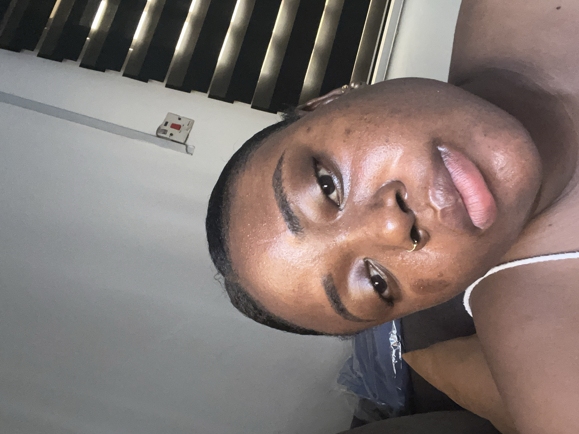 A close-up, angled shot features a dark-skinned person with a visible nose ring and subtle makeup. The person is facing slightly downward toward the camera.
In the background, there is a partially visible window covered by blinds, and a white electrical outlet is set against a pale gray wall.