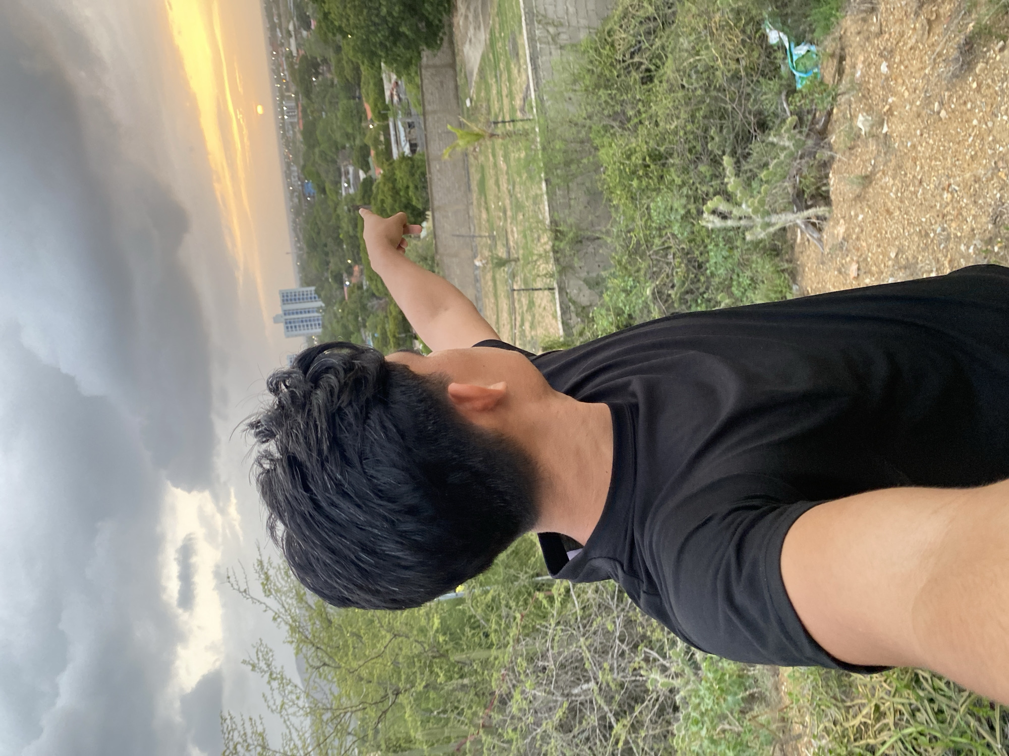 In Juan Griego, Venezuela, a person wearing a black t-shirt stands on a hillside, pointing towards the horizon where a sunset is visible. The sky is partially overcast with dark clouds, but a bright orange glow emanates from the setting sun, illuminating a distant city skyline. The foreground is dominated by dry brush and sparse vegetation, with a concrete wall visible in the mid-ground. The person's face is not visible, as they are turned away from the camera, looking out at the landscape. The image captures a moment of contemplation or discovery, with the natural beauty of the sunset juxtaposed against the urban environment.
