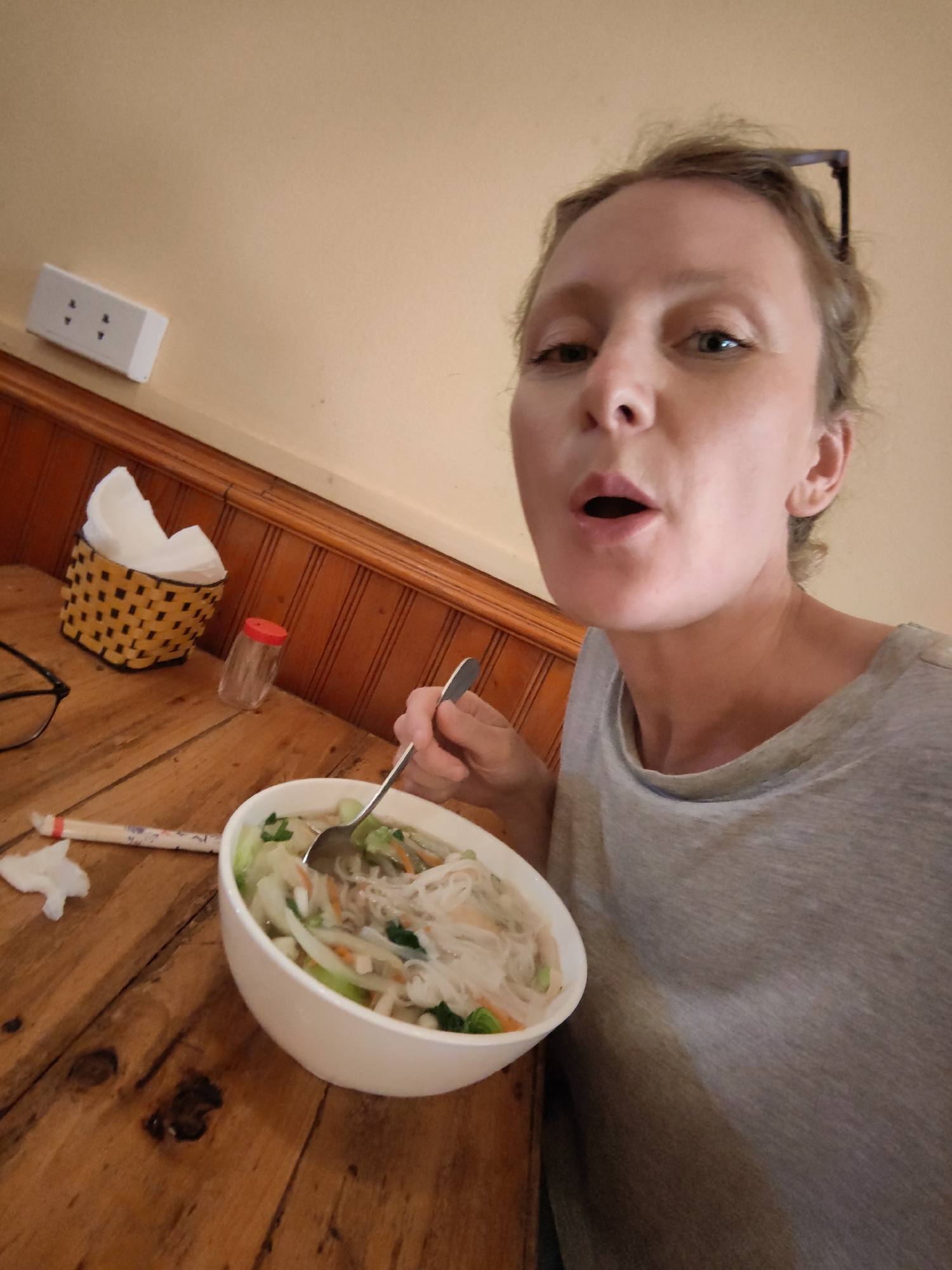 A woman with light-colored hair and a casual grey t-shirt is captured in a close-up selfie or photo, appearing to be in the middle of eating. Her mouth is slightly agape, suggesting she is about to speak or take a bite, and her eyes are looking directly towards the camera. A pair of sunglasses is perched on top of her head.
She is seated at a wooden table with a visible grain. In front of her is a large white bowl filled with what appears to be a noodle soup, possibly a Vietnamese dish like pho or bun, containing white vermicelli noodles, sliced carrots, green leafy vegetables, and onions in a clear broth. She holds a spoon in her right hand, immersed in the bowl.
On the table to her left, there's a pair of black-rimmed eyeglasses. Beside them, a crumpled white napkin or tissue rests near a pair of chopsticks enclosed in a white paper wrapper with faint red and blue markings. Further back on the table, a yellow and black woven basket holds a stack of white paper napkins, and next to it, a small, clear shaker with a red cap is visible, likely containing a condiment.
The background features a wall composed of two distinct sections: a lower portion with dark wooden paneling, characterized by vertical slats, and an upper portion painted a uniform light beige or pale yellow color. On this upper wall, a white electrical outlet with two sets of two-pronged sockets is mounted. The overall lighting appears soft and natural, suggesting an indoor setting during the daytime.