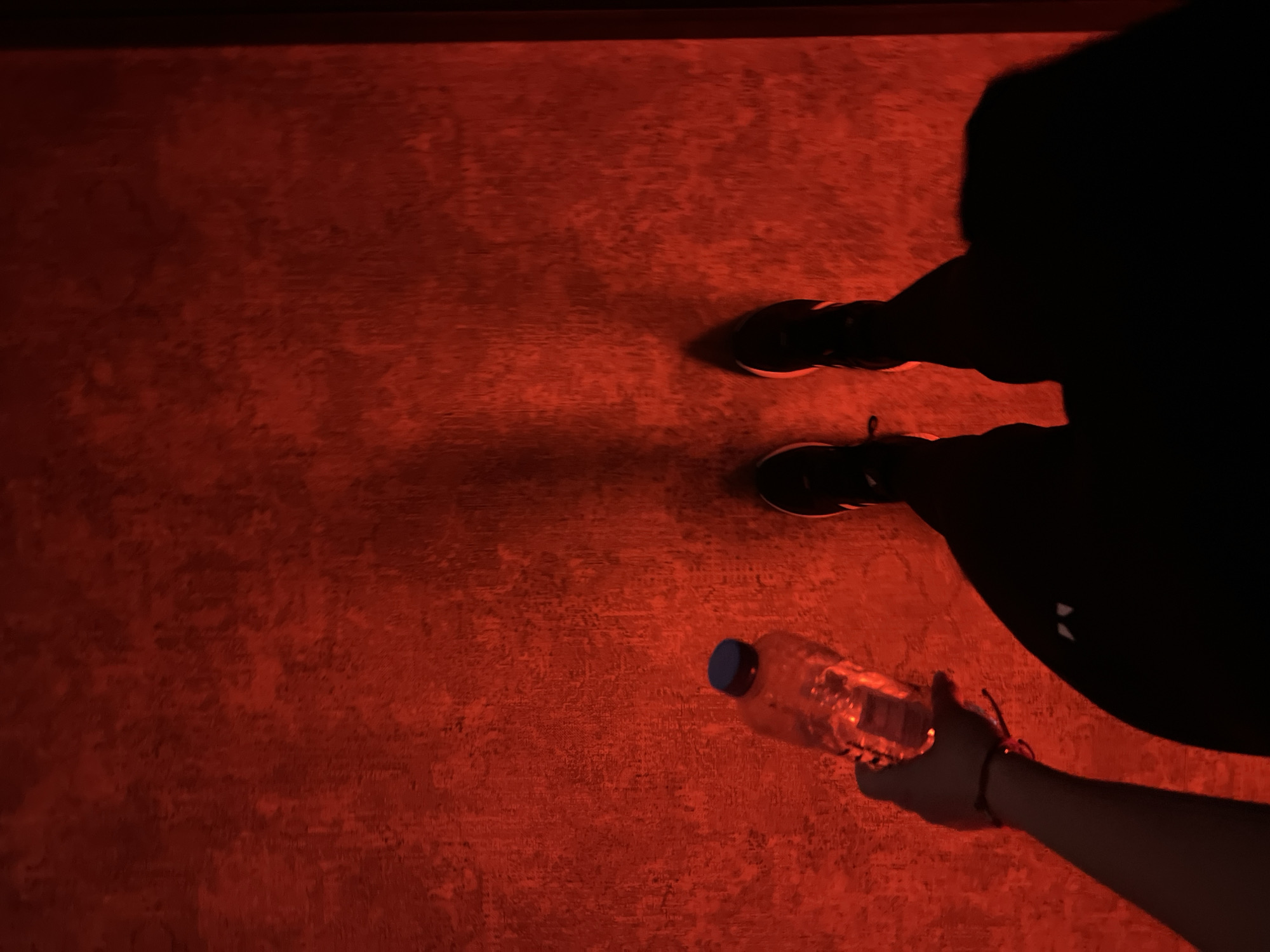The scene is indoors, under dim red lighting. The floor is textured and appears to be concrete. A dark silhouette of a person stands on the right side of the image, wearing shoes. A hand and arm reach into the scene from the lower right corner, holding a plastic water bottle. The location is unknown, and its relation to Plovdiv, Bulgaria is unclear.