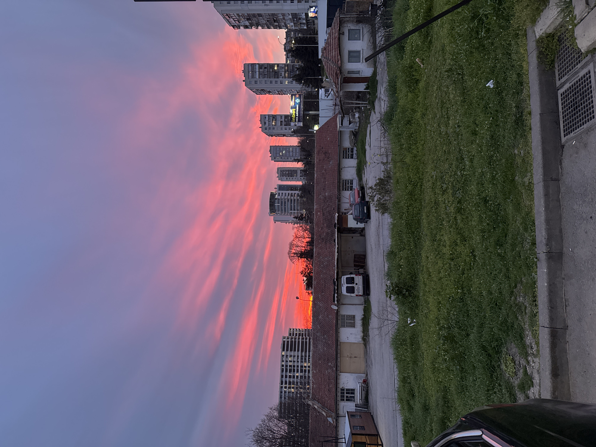 The image shows a view of Varna, Bulgaria at sunset. The sky displays vibrant hues of pink and purple with streaky cloud formations. Multiple high-rise apartment buildings are visible in the background, interspersed with trees. In the foreground, there is a row of older, single-story buildings with reddish tile roofs. A narrow street runs between the buildings and a grassy area, bordered by a concrete curb. A white van is parked along the side of the street. A car is partially visible in the lower right corner of the image. The photo is rotated ninety degrees clockwise, so the sky appears on the left and the ground on the right.