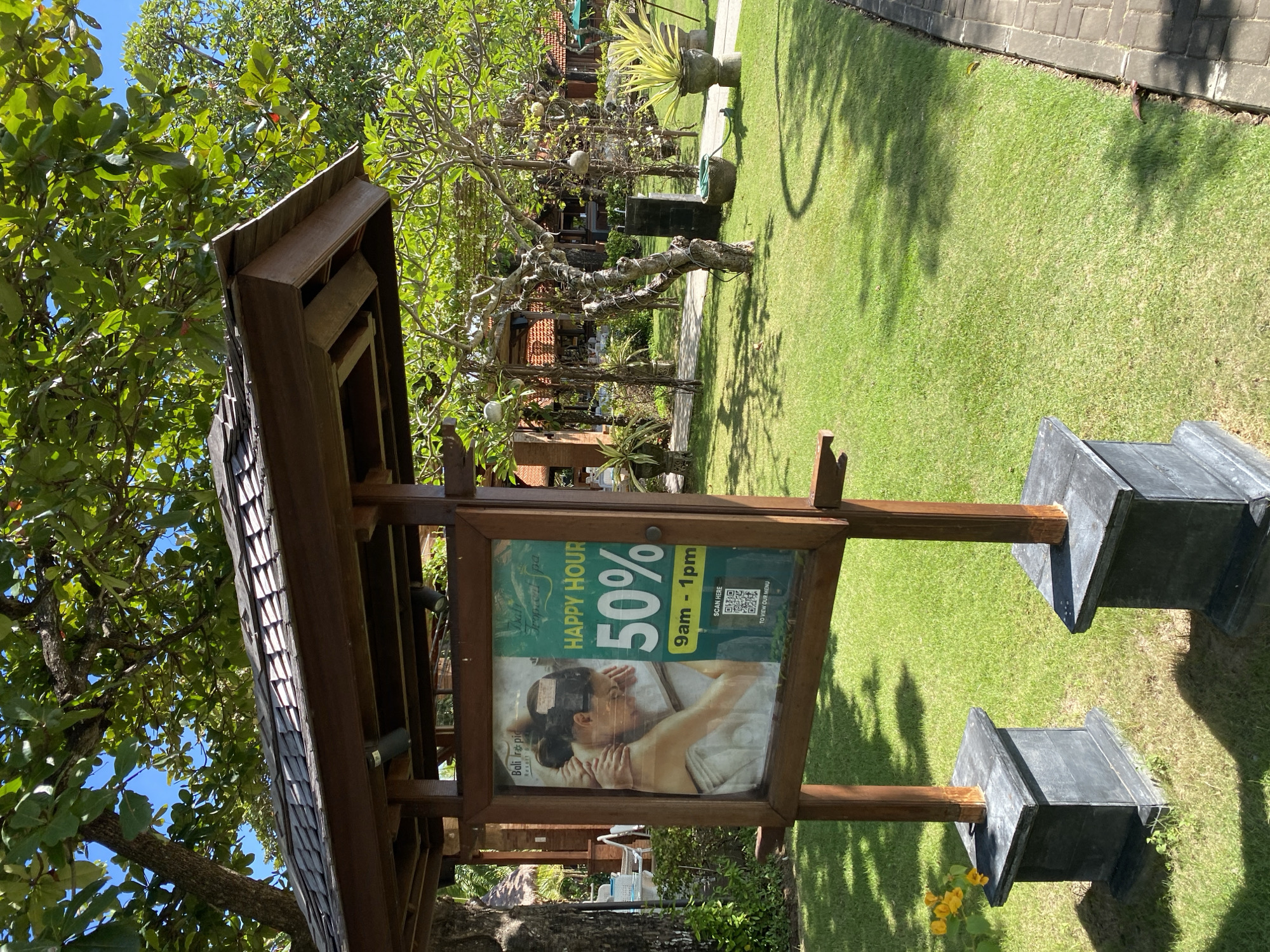 The image captures a wooden kiosk situated on a well-maintained lawn. The kiosk displays a sign promoting a "Happy Hour" at "Bali Irs pic Resort B", offering a 50% discount from 9 am to 1 pm. A QR code is also visible on the sign, offering a link to view the menu. The scene is taken in an outdoor setting with green grass covering the ground. The presence of trees and other vegetation suggests the location is likely a resort or spa in Kuta Selatan, Indonesia.