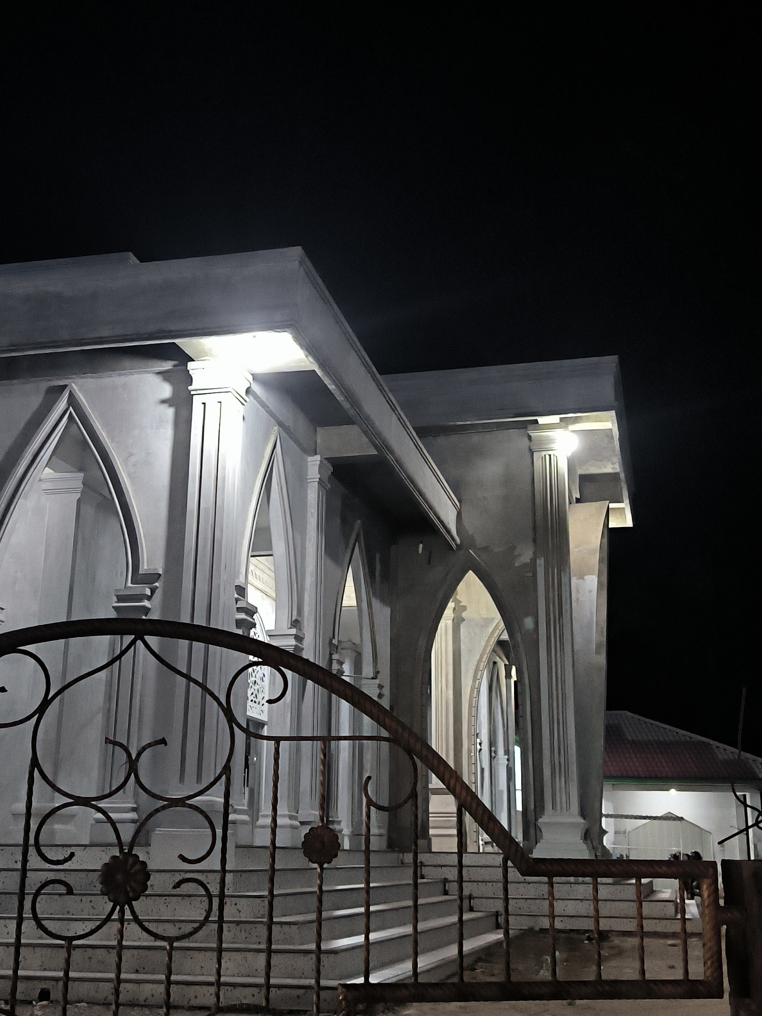 An eye-level, outdoor photograph captures a gray structure with multiple arches and columns at night in Alueraya, Indonesia. A wrought iron gate with floral designs in the lower portion and curling designs above stands in the foreground, partially obscuring the view. The structure's architecture features several arches and columns, creating a rhythmic, repeating pattern. Lights are visible above each column, illuminating the area. Behind the main structure, a building with a red roof is visible. The sky is a dark, indistinct background. The scene is still, suggesting it was taken when there was little activity.