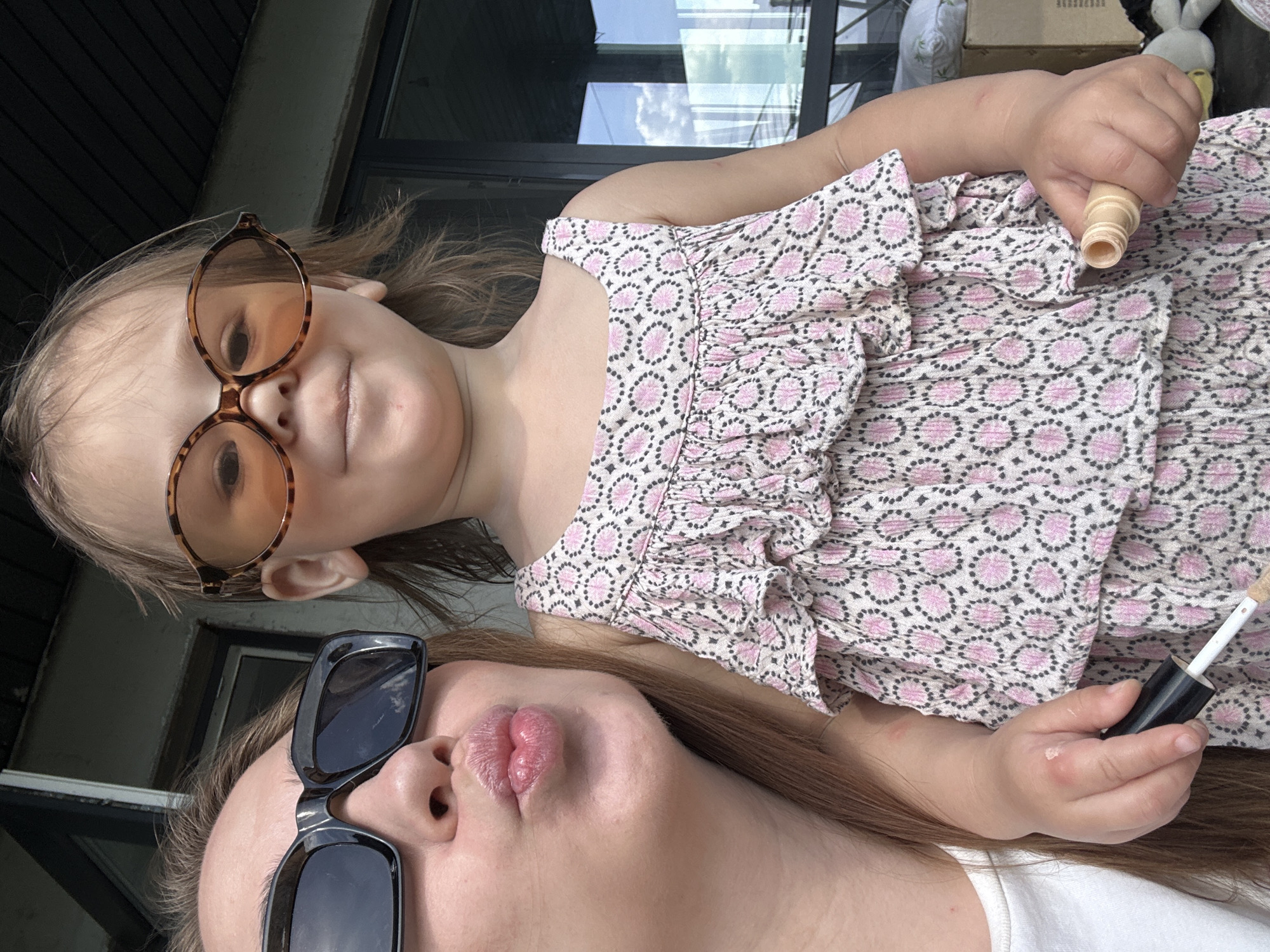 A woman and a young child are positioned close together in what appears to be an outdoor setting. The woman is wearing sunglasses and pursing her lips toward the camera. The child, also wearing sunglasses, is holding makeup and looking at the camera. The background shows an exterior with windows. There is no definitive indication of Kharnavy, Russia.