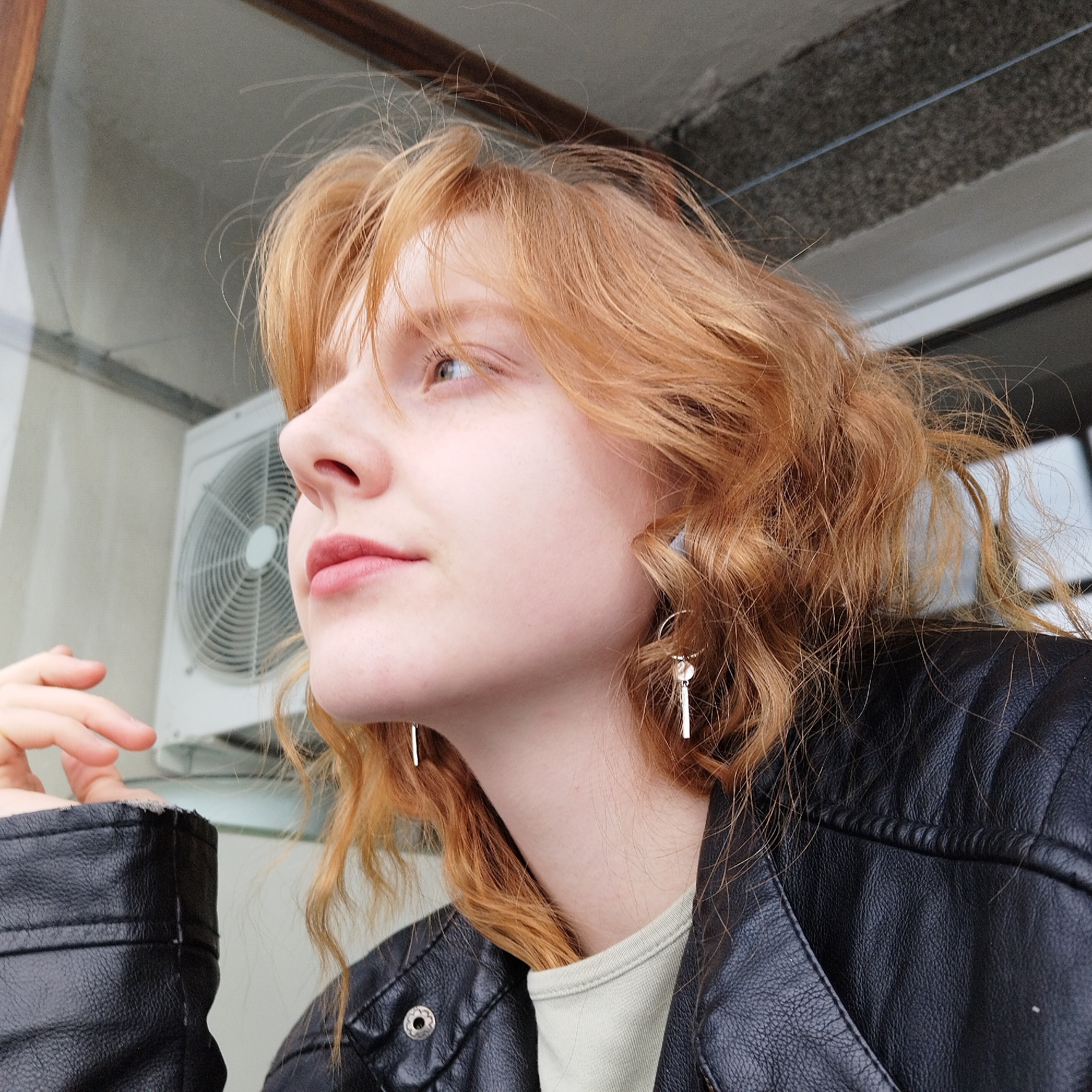 A fair-skinned young woman with strawberry blonde hair is gazing leftward, possibly outside of a window. She is wearing a black leather jacket and light-colored t-shirt. She has thin gold earrings, and her hand is partially visible near the window frame. An air conditioning unit is visible outside the window, and what seems to be a concrete balcony or ledge. It is not possible to determine if this image was taken in Yekaterinburg, Russia, from the details provided.