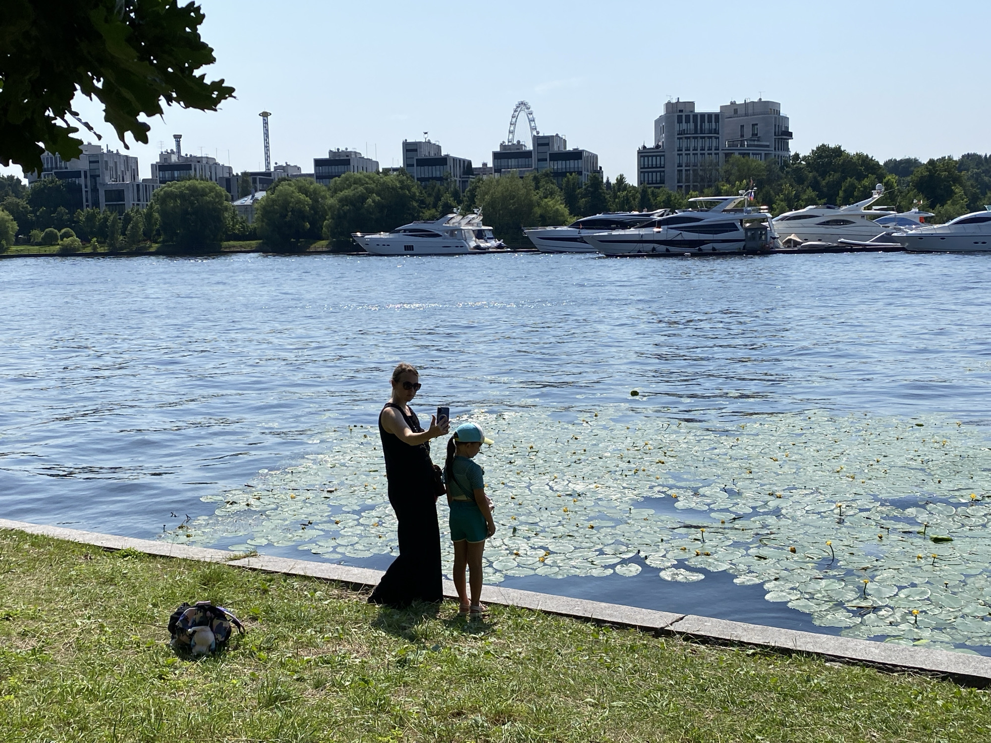 A woman in a long black dress and sunglasses stands on a grassy bank next to a young girl wearing a blue cap and green shorts. The woman is holding a smartphone, appearing to take a picture or video of the girl and the water. The girl is looking towards a body of water, which is covered in a dense carpet of lily pads with small yellow flowers. Behind the water, a row of modern, multi-story buildings and trees are visible, suggesting an urban waterfront. Several large yachts are docked in the water. In the far background, a Ferris wheel and tall structures are discernible against a clear blue sky. A patterned bag rests on the grass to the left of the two figures. The scene appears to be a leisure area along the Neva River or one of its tributaries in Saint Petersburg, Russia, possibly near a yacht club or waterfront development.