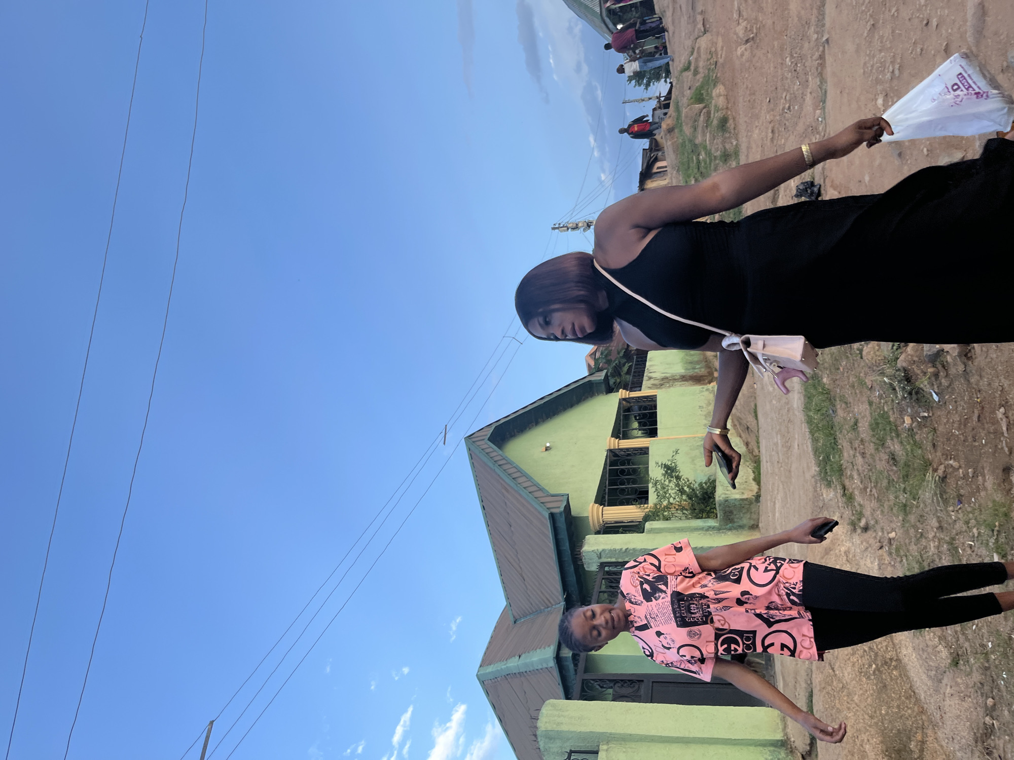 Two young women are depicted outdoors in a setting that appears to be a residential area in Jos, Nigeria. The image is vertically oriented and shows a building with a pale green facade and a dark green roof. Power lines are visible against a clear blue sky with scattered white clouds, suggesting it is daytime, likely late afternoon due to the soft lighting.
In the foreground, one woman is wearing a pink t-shirt with a repeating pattern of black text and logos, including what appears to be "GUCCI", and black leggings. She is looking downwards with a slight smile.
In the background, slightly above and to the right of the first woman, another woman is dressed in a black sleeveless dress. She is holding a white plastic bag in her right hand and has a small, light-colored purse slung across her shoulder. Her left hand is reaching towards her right side.
The ground surface is unpaved and appears to be made of dirt and gravel, with some debris scattered around. In the far background, more buildings and several people are visible, hinting at a populated area. The overall mood of the image is casual and relaxed, capturing a moment in everyday life.