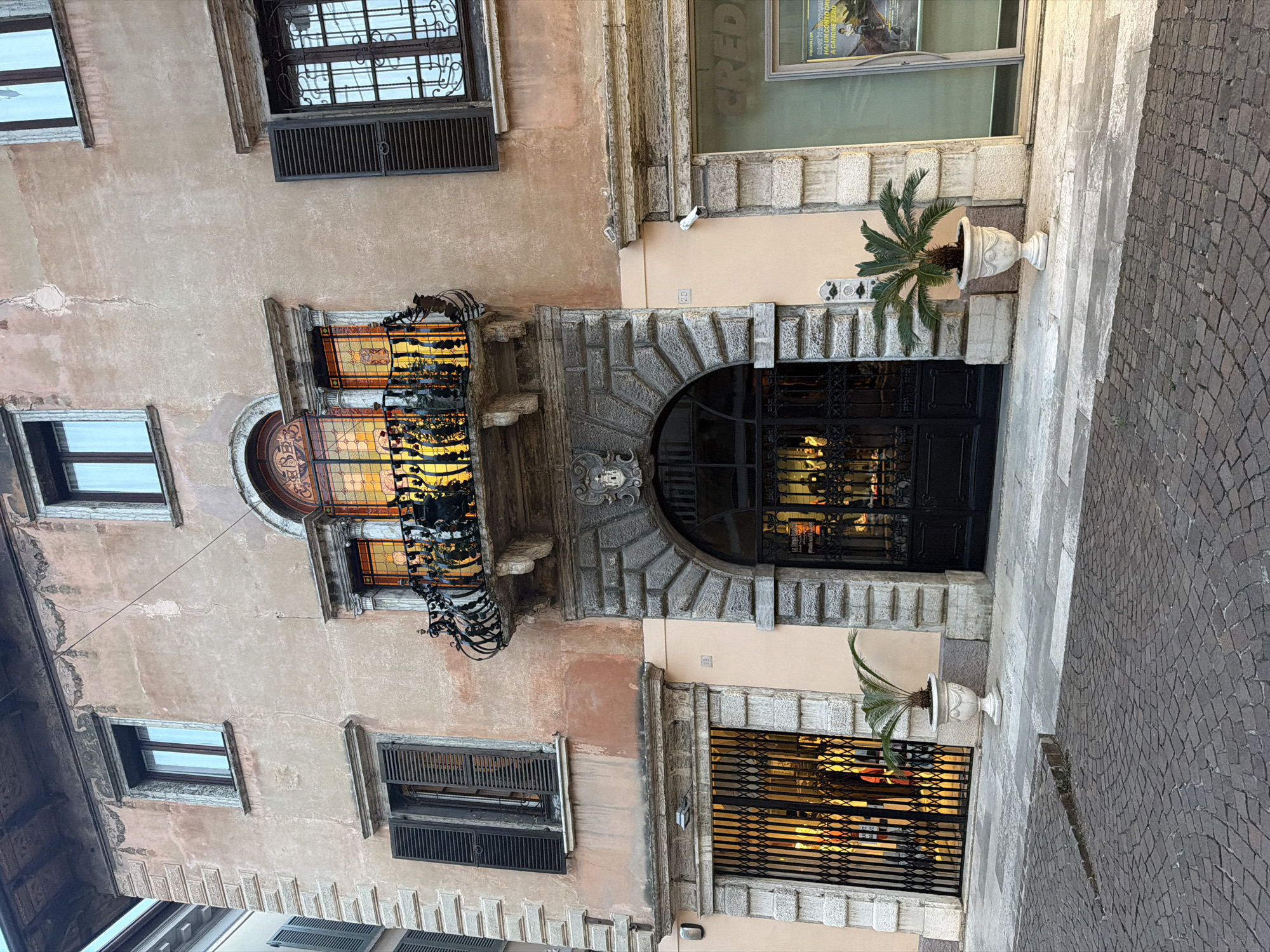 The image shows the facade of a multi-story building in Salò, Italy. The building has a pinkish-ochre colored plaster facade with decorative elements around the windows and balconies. There are several windows with dark shutters. The main entrance is a large arched doorway with intricate stone detailing and a black metal gate. Above the entrance, there is a balcony with a decorative wrought-iron railing and stained-glass panels in an arched window. To the left of the main entrance is a shop with a metal security gate, and to the right is a storefront with a large glass window and signage for "CREDI". Potted palm plants are placed on the pavement in front of the building. The pavement is paved with cobblestones. The scene appears to be on a street in a European city. There are no people visible in the image. The lighting suggests it is daytime.
