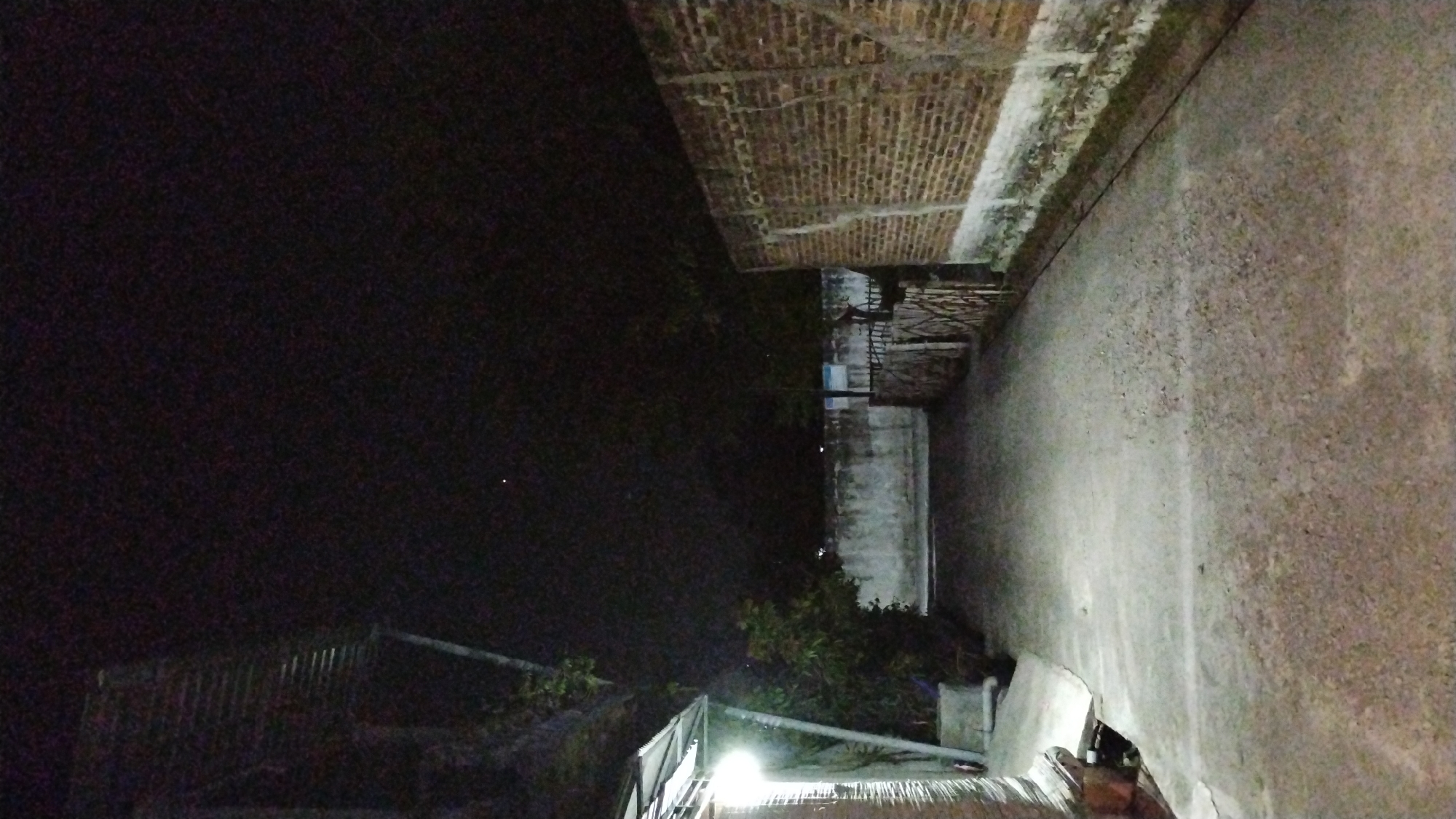 The image depicts a dimly lit pathway at night in Banjarsari, Indonesia. The pathway is paved with a textured, light-colored material and is flanked by walls on both sides. On the left, there is a building with a corrugated metal roof extending over the pathway, illuminated by a bright light source. Some foliage is visible near the building. On the right side, a wall made of reddish-brown bricks runs along the pathway. The wall has a weathered appearance with white paint or plaster at its base. Further down the pathway on the right, a wooden fence is visible. The sky above is dark, suggesting nighttime, and two small, distant points of light, possibly stars or distant lights, are faintly visible in the sky. The overall atmosphere is one of quietness and low light.