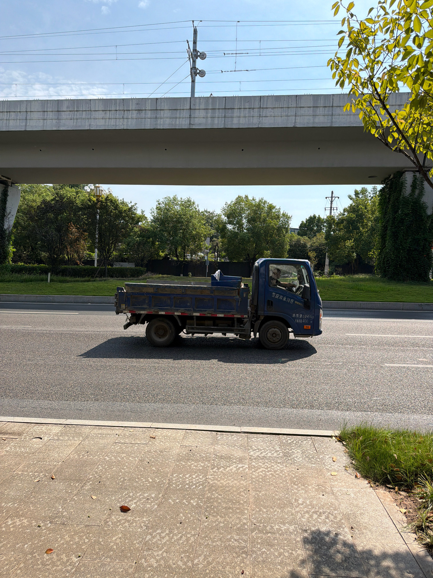 An exterior, eye-level, medium shot shows a blue dump truck driving on a paved road under an overpass in Jiangjin, China. The truck is facing right and is partially shaded by the overpass. The driver is visible in the cab. The overpass is made of concrete and has overhead wires. Trees and foliage are visible behind the road. The foreground features a sidewalk with scattered leaves. The sky is blue with some clouds.