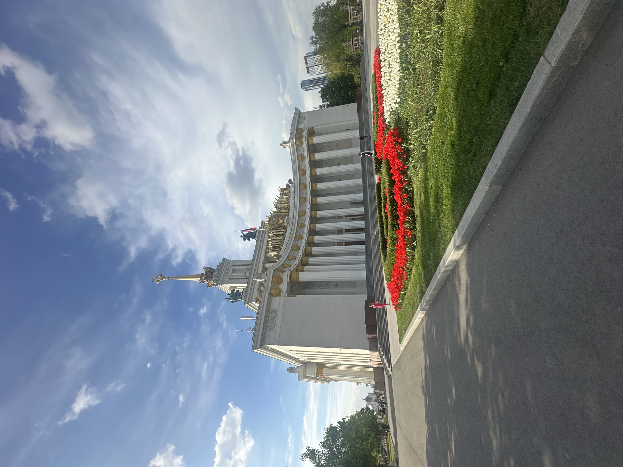 The image captures a building in the VDNH (Exhibition of Achievements of National Economy) area of Moscow, Russia. The building is white with gold architectural details and a spire. Red and white flowers are planted next to the building, separated by a strip of green grass. The sky is blue with white clouds. A paved walkway is visible in the foreground. A person in a red dress can be seen walking up the steps of the building. In the background, buildings and trees are visible.