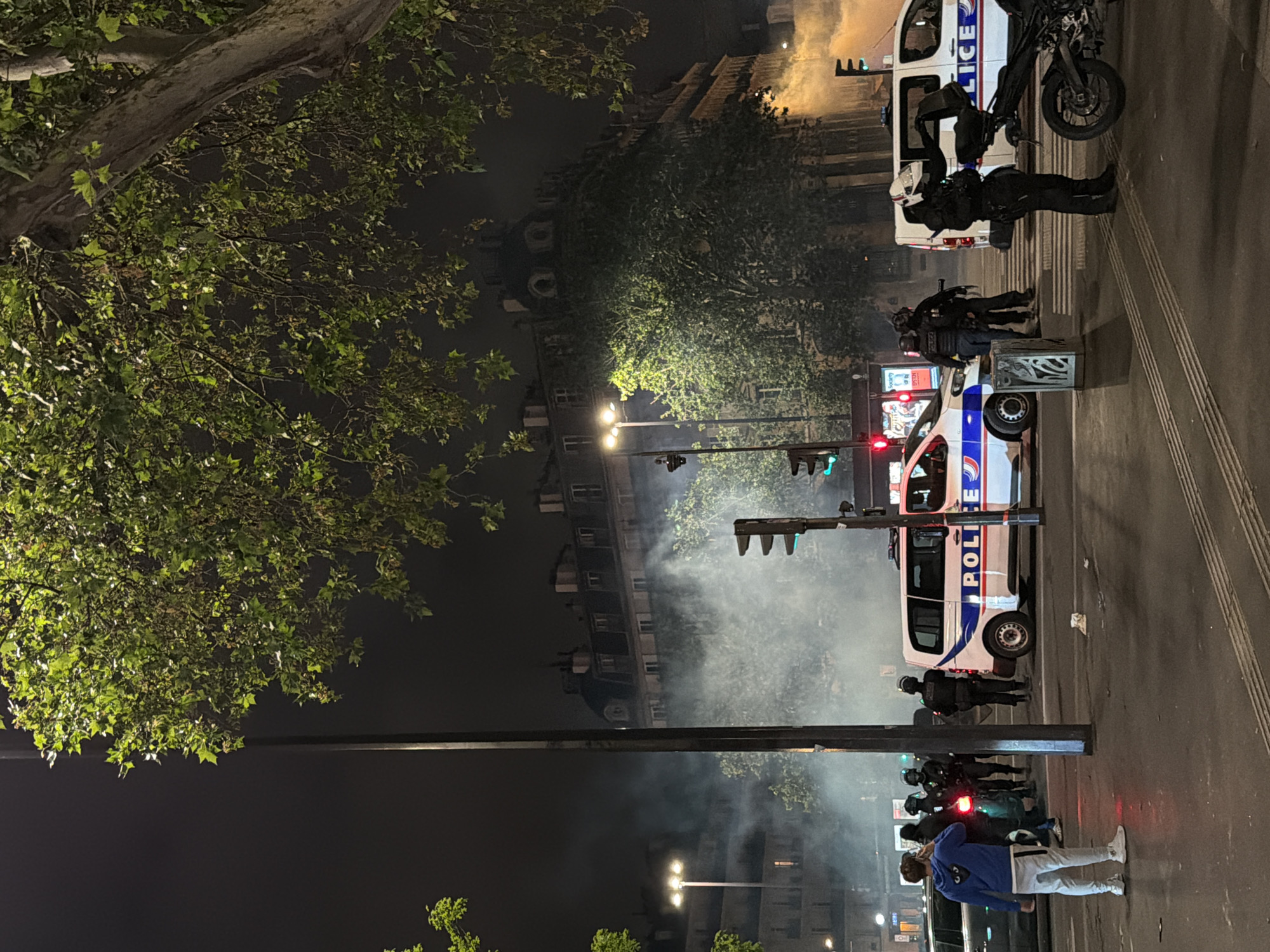 The image depicts a nighttime scene in Paris, France, likely during a protest or unrest. Several police vehicles are present, with officers in riot gear standing nearby. Smoke fills the air, obscuring the background buildings. A few civilians are visible, including one in a blue jacket and light-colored pants standing on the sidewalk. The overall atmosphere is tense and suggestive of conflict. Traffic lights are visible in the street, indicating the scene is on a street or avenue. The presence of police vehicles marked "POLICE" confirms the location is likely France.