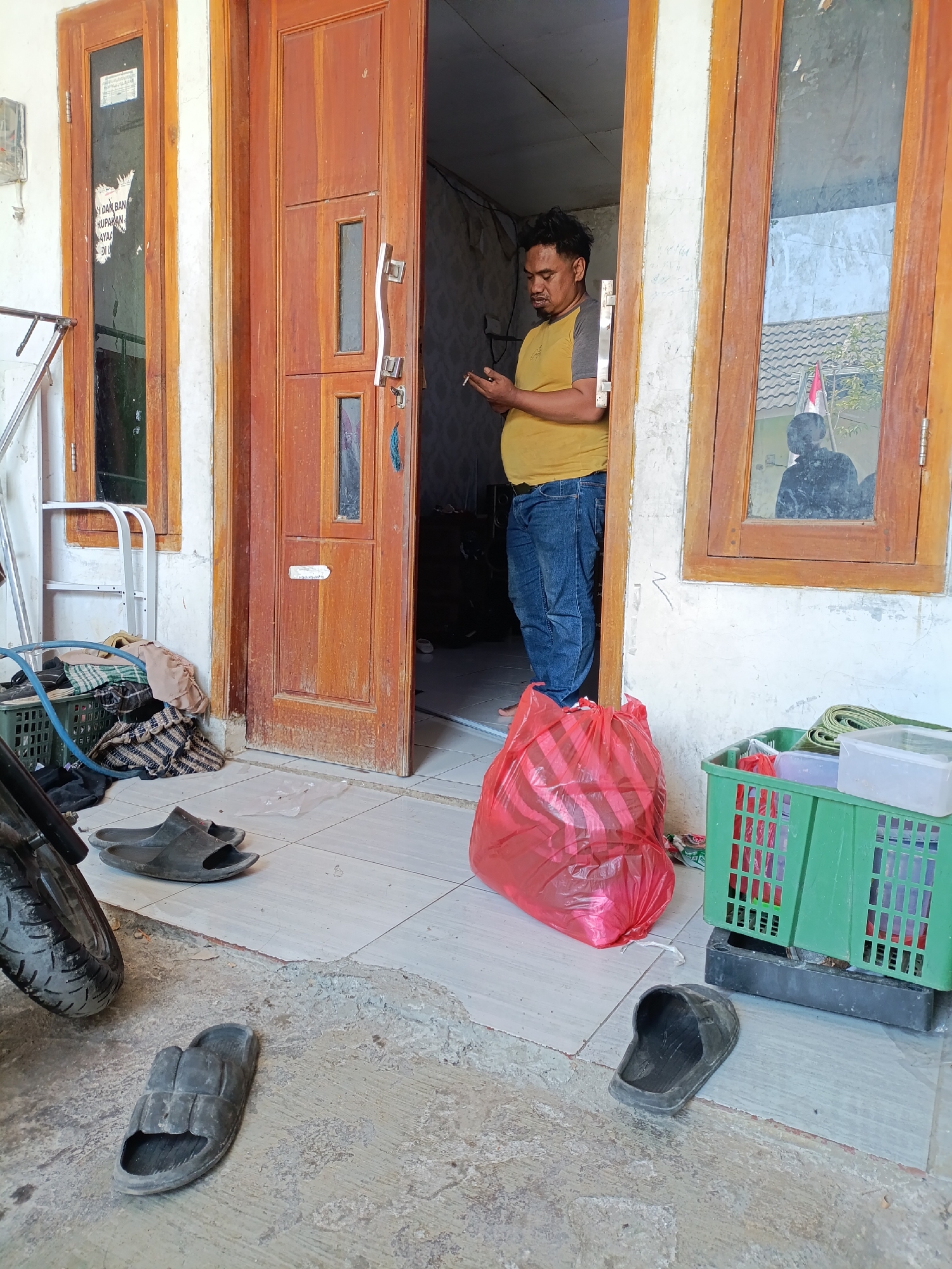 An Indonesian man is standing in the doorway of a house in Cariu, Indonesia. He is wearing a yellow t-shirt and blue jeans and is smoking a cigarette. He is looking at his phone. To his left is a wooden door with two windows and a metal handle. To his right, a large red plastic bag is on the floor. A green plastic crate with some items inside is next to the bag. Several pairs of black flip-flops are scattered on the tiled floor in front of the house. A motorcycle wheel is visible in the lower left corner of the image. The wall of the house is white and somewhat weathered.