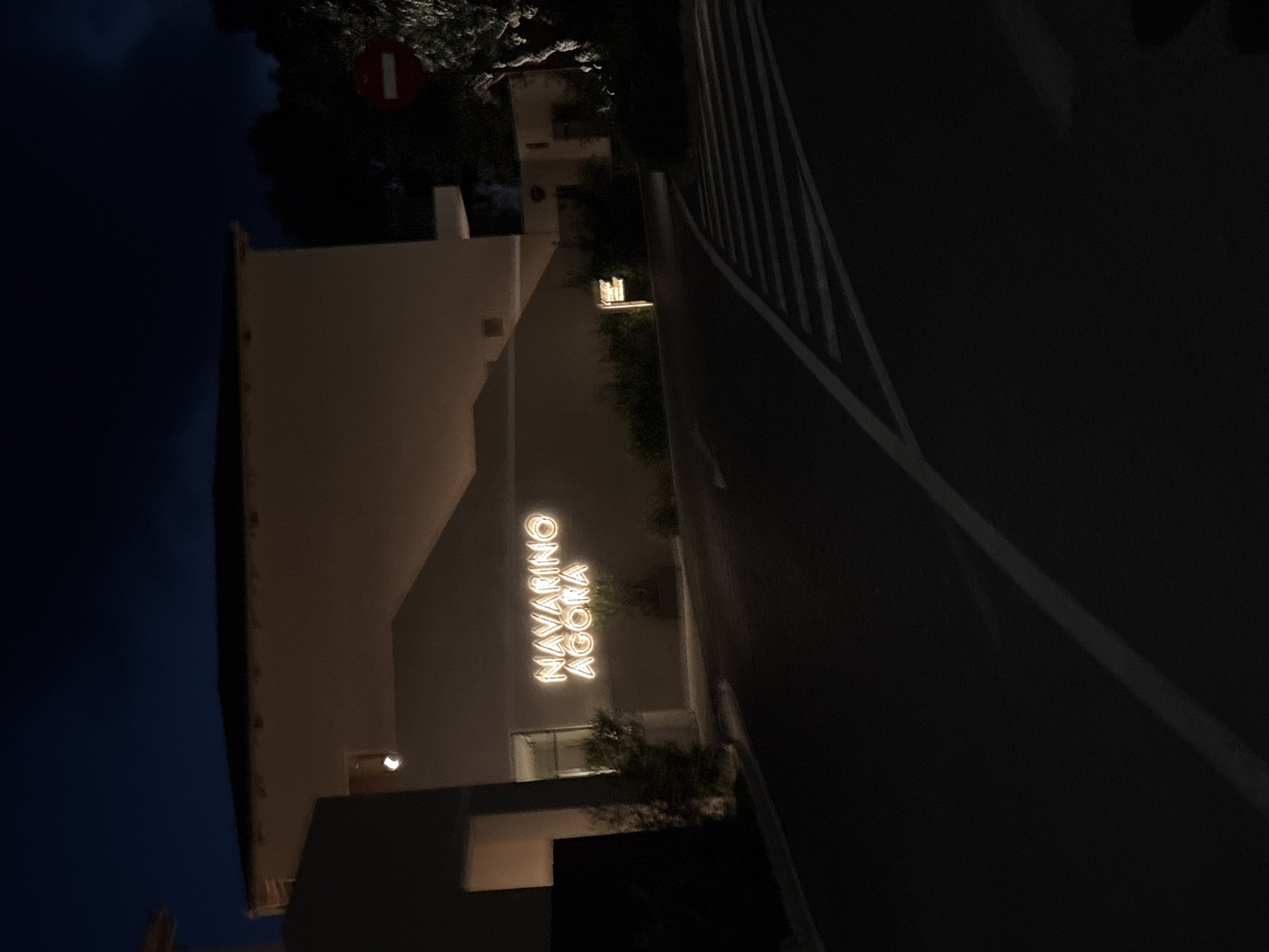 The image captures an outdoor scene at night, featuring a modern building with an illuminated sign and an adjacent paved area or road. The dark sky indicates late evening or nighttime.
On the left, a light-colored building is visible, with a prominent, vertically oriented sign illuminated in a warm, golden light. The text on the sign clearly reads "NAVARINO AGORA" in capital letters. Subtle lighting also accentuates parts of the building's facade and surrounding vegetation, which appears as dark foliage.
To the right of the building, a dark, paved surface, likely a road or a wide pathway, stretches into the background. White lines are visible on this surface, suggesting lane markings or boundaries. Additional dark vegetation and trees are scattered around the building and along the edges of the paved area. In the upper left portion of the image, partially obscured by dark leaves, a red circular sign with a white horizontal bar, typically denoting a "do not enter" restriction, can be seen.
No people or active events are present in the image, conveying a sense of quiet and stillness. The scene appears well-maintained, highlighting the "Navarino Agora" as a focal point, likely a commercial or public area in Pylos, Greece.