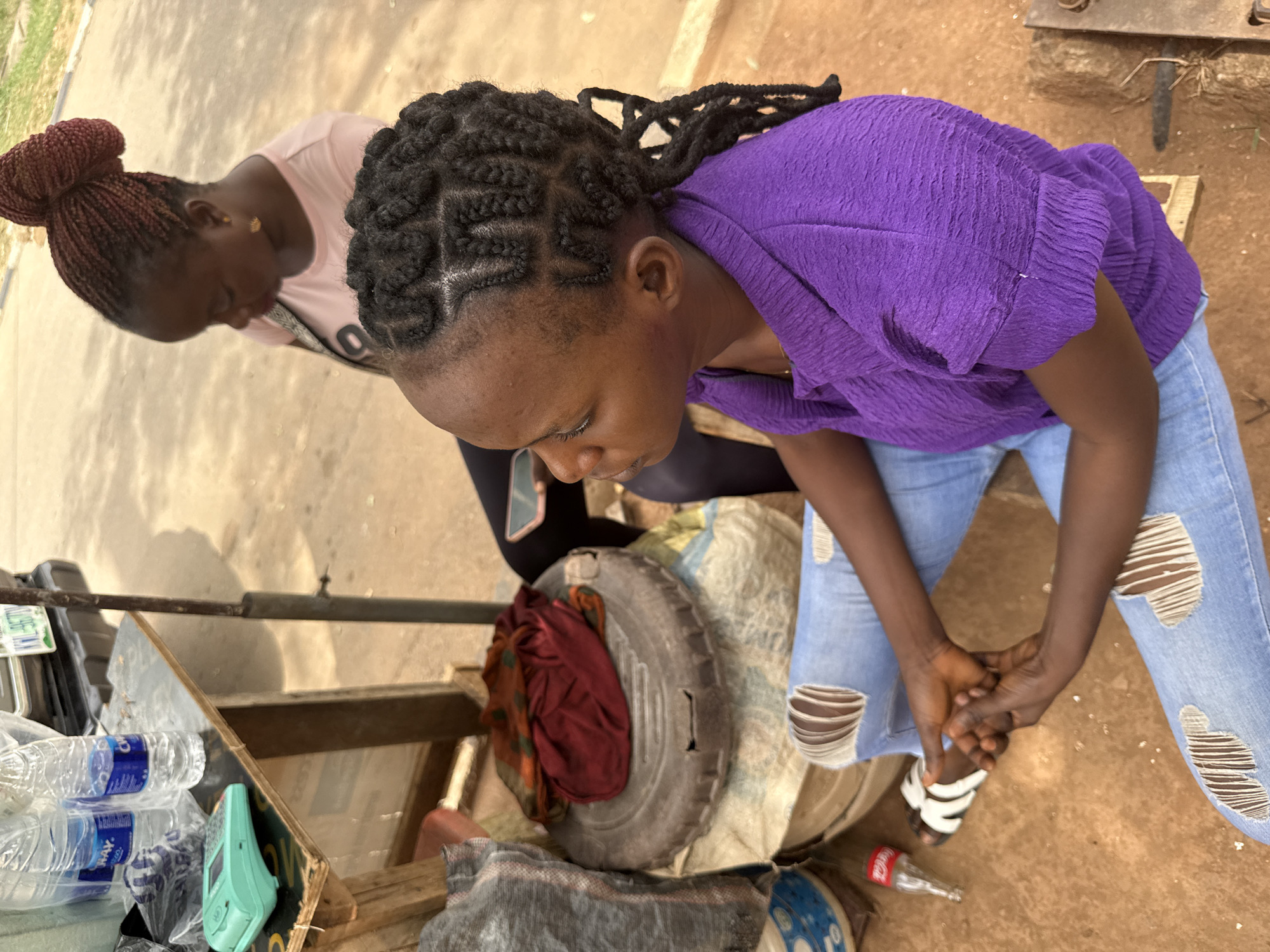 Two women are outdoors in Abuja, Nigeria. One woman wears a purple shirt and ripped blue jeans. The other woman wears a pink shirt and dark pants. Both women have braided hairstyles. They stand near a table laden with items, including water bottles and a spare tire. The woman in the purple shirt has her hands clasped in front of her.