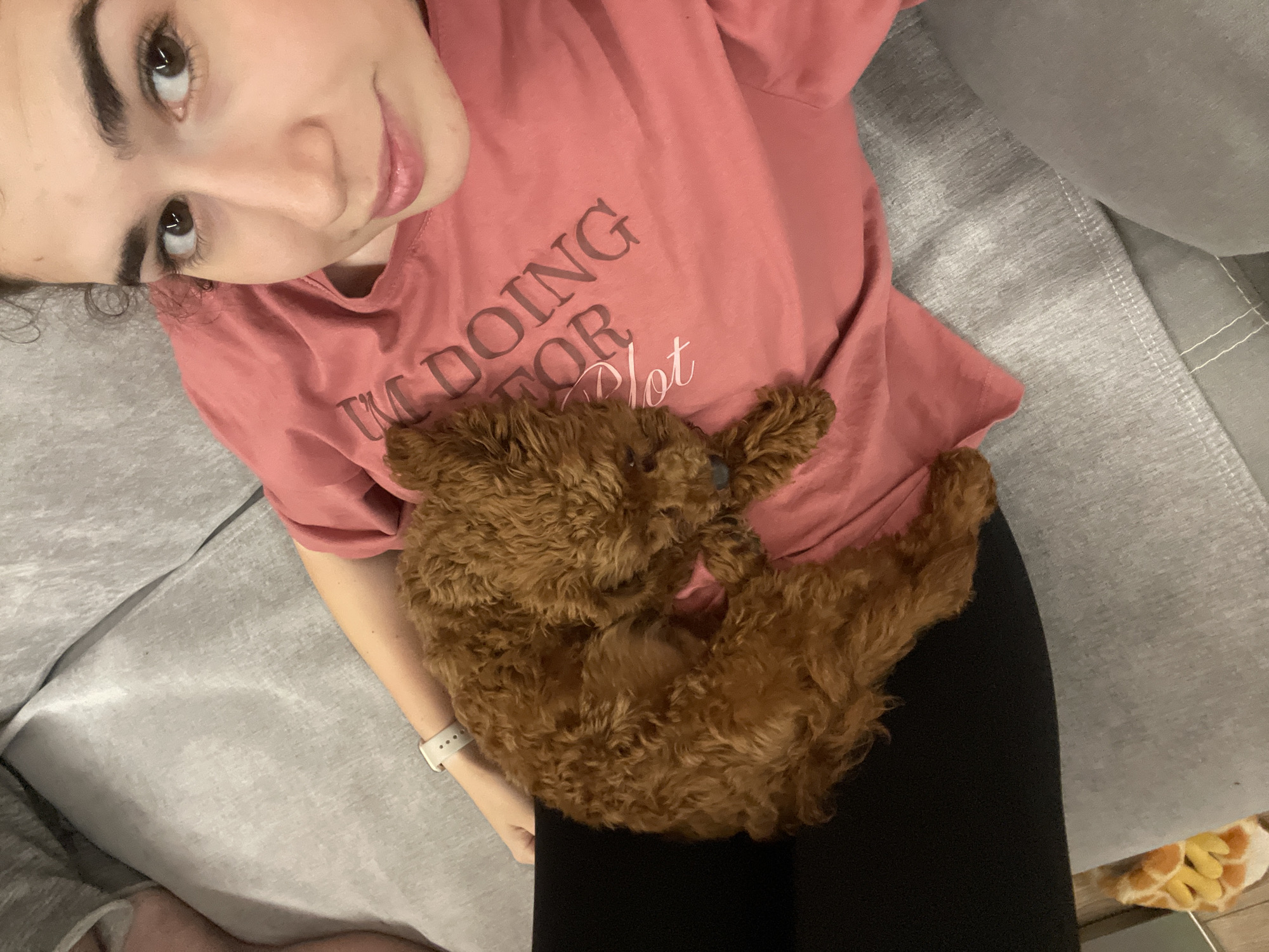 A woman is lying on a gray couch and looking at the camera. She has dark hair and is wearing a pink t-shirt with text that is only partially visible: "I'M DOING FOR Plot". She is also wearing a white smartwatch. A small, brown, curly-haired dog is lying on her chest. The woman is wearing black leggings. The location is not specified in the image and cannot be determined as Sofia, Bulgaria.