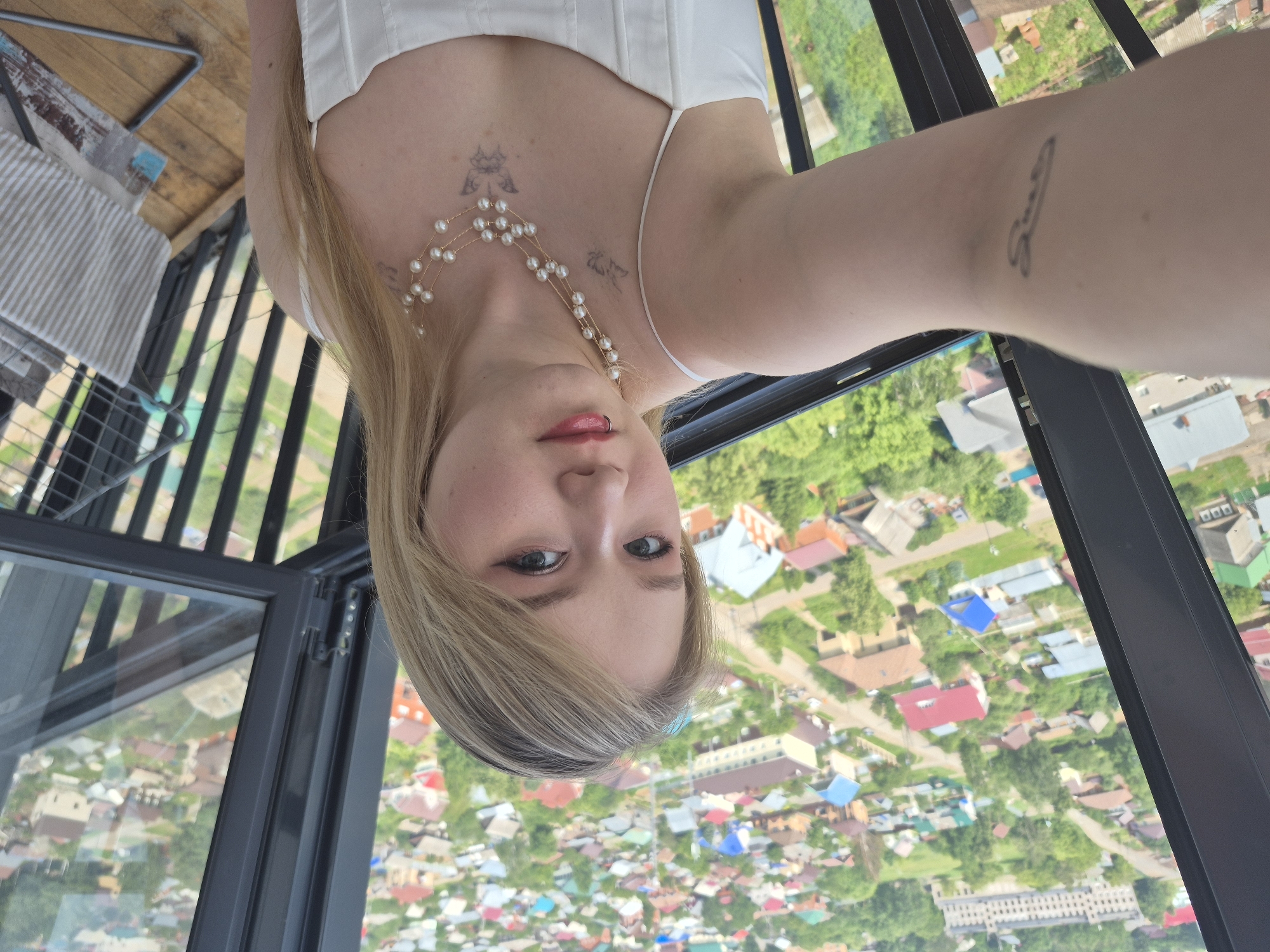 A young woman with blonde hair and makeup is taking a selfie on a balcony overlooking a residential area in Izhevsk, Russia. She is wearing a white dress and a pearl necklace. There are tattoos on her arm and chest. The background shows numerous houses with colorful roofs, trees, and a large building in the distance. The balcony has metal railings and a wooden floor. A clothes drying rack with striped towels is visible on the right side of the image. The overall impression is a snapshot of a person enjoying a scenic view from an elevated position.