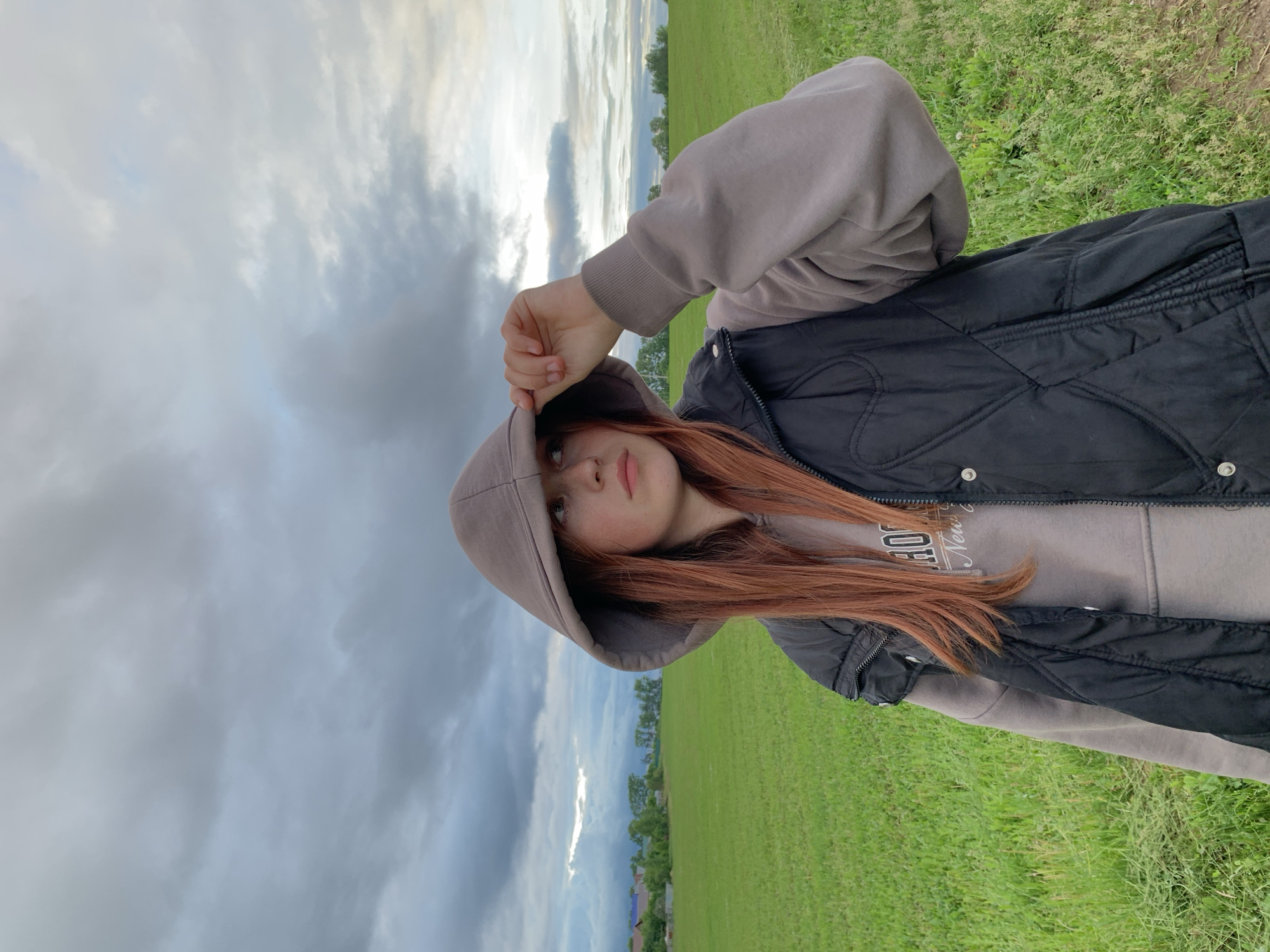 A young individual is captured in a waist-up outdoor portrait in Фоки, Russia. The subject, a female with long, reddish-brown hair, is wearing a light brown hooded sweatshirt with the hood drawn over her head and a black quilted sleeveless vest over it. Her left hand is raised, adjusting the front edge of the hood. She has light skin, visible freckles, and is looking off-camera to the viewer's right with a neutral expression. The background features an expansive, vibrant green field under a heavily clouded, gray sky with hints of brighter, possibly sunlit areas towards the upper right horizon. In the distant left, a cluster of houses or buildings with varying roof colors is visible amidst trees, suggesting a rural settlement. The overall scene is illuminated by soft, diffused light.
