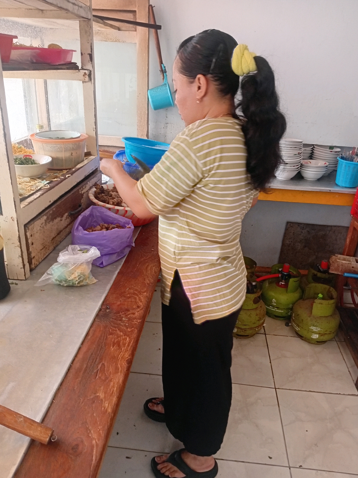 The image depicts a woman standing behind a food stall in Besito, Indonesia. She is wearing a yellow and white striped t-shirt and black pants, with her dark hair tied back in a ponytail secured by a yellow scrunchie. Her hands are engaged in preparing food, with one hand holding a spoon and the other near a bowl of ingredients.
The stall features a wooden counter and shelves displaying various food items and cooking utensils. To the left, there are red and orange bowls containing noodles and other food. In front of the woman, there are several bags, including a purple bag filled with what appears to be meat and a clear plastic bag containing green vegetables. Behind her, stacks of white bowls are visible on a shelf, along with a blue basket holding utensils and several green gas cylinders on the floor. The overall environment suggests a busy food preparation area.