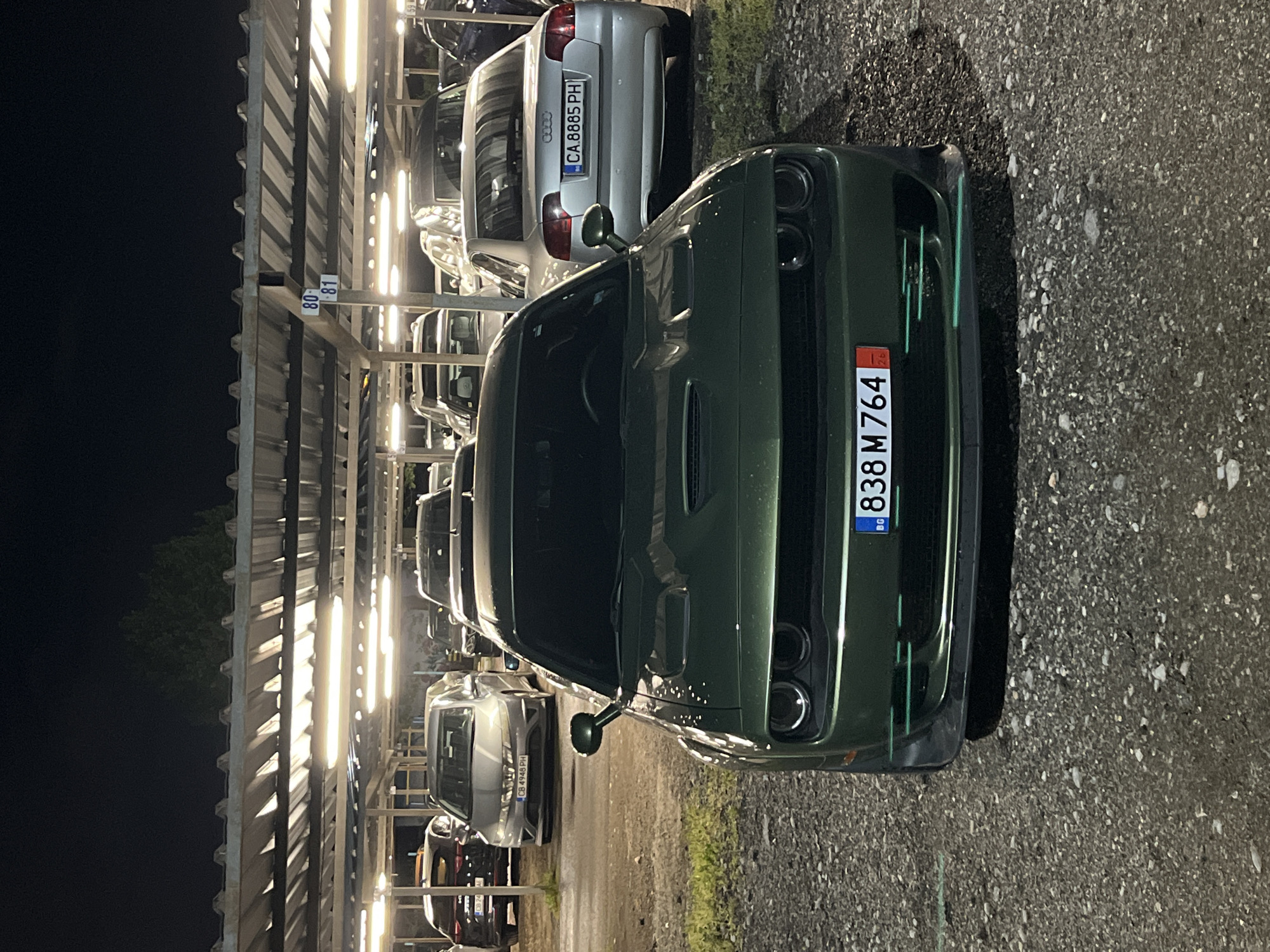 The image displays a nighttime scene within what appears to be a multi-level or covered outdoor parking facility in Sofia, Bulgaria. Rows of bright, artificial lights illuminate the area, highlighting the parked vehicles and the metallic support structure.
The primary subject is a dark green Dodge Challenger, prominently parked in the foreground on a gravel and asphalt surface. Its front fascia, including a hood scoop and distinctive quad headlights, is clearly visible. The car's license plate, located on the front bumper, reads "BG 838 M 764".
Behind the green Challenger, a silver Audi sedan is parked, facing away from the camera. Its rear license plate, "CA 8885 PH", is fully legible. Further into the background, other cars are visible, including another silver vehicle with a partially visible license plate "CB 4948 PH".
The parking structure consists of metal beams and corrugated panels. On one of the vertical supports, two parking bay numbers, "80" and "81", are displayed. The left side of the image shows the dark night sky, contrasting with the brightly lit parking area. No people are present, and the scene depicts a static arrangement of parked automobiles.