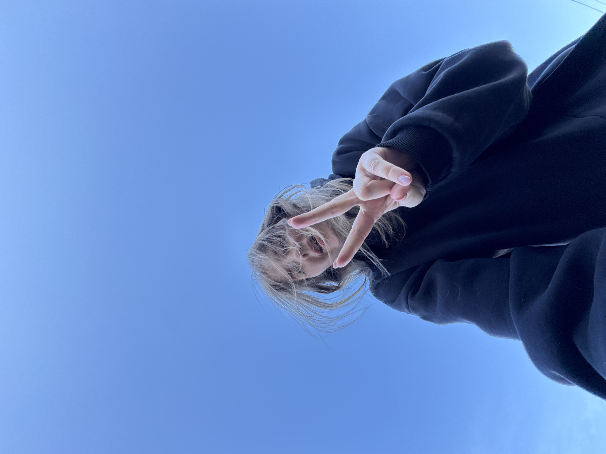 A low-angle, upward-facing photograph depicts a person with fair hair wearing a dark hooded sweatshirt against a backdrop of clear blue sky. The person is making a peace sign with their right hand, held aloft. The subject's face is visible, partially obscured by strands of hair. The location could be Ферма, Russia, based on user request. The photograph is angled, tilting the subject to the right.