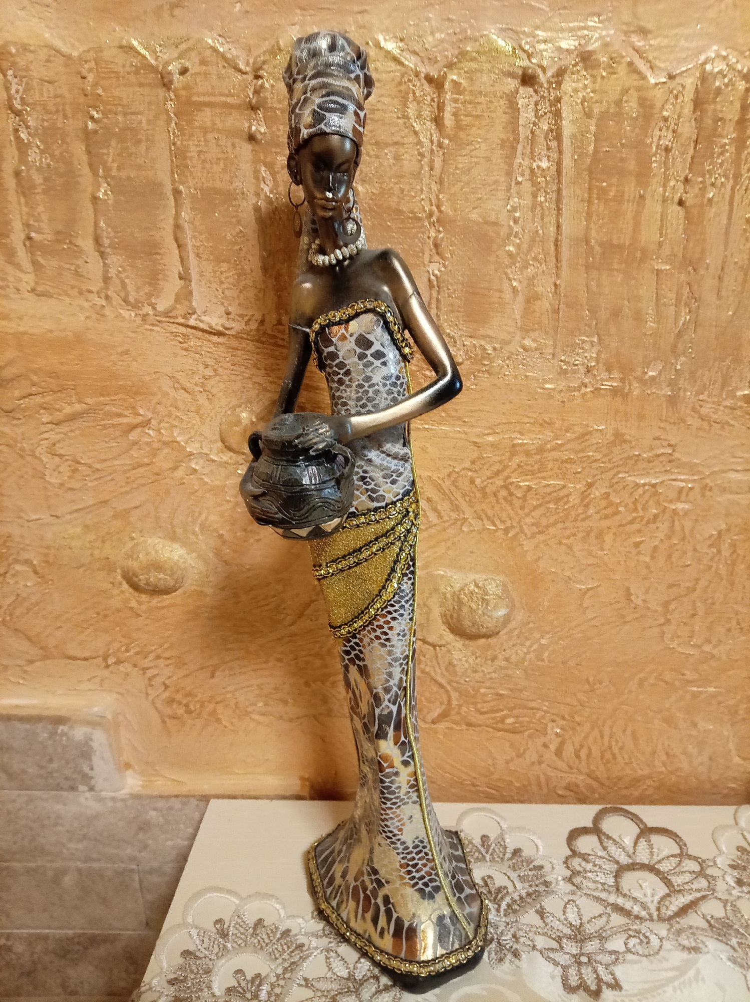 An ornate figurine of an African woman stands on a lace-covered surface, set against a textured, golden-brown wall with vertical ridges. The figurine is depicted wearing a patterned dress with gold accents, a pearl necklace, and earrings. She holds a dark, textured pot in her left arm. The background wall has raised circular elements and a rough, stucco-like texture. The foreground shows a light-colored surface adorned with intricate white lace patterns. The lighting is warm, highlighting the metallic sheen of the figurine and the textured details of the wall. The scene appears to be an indoor still life.
