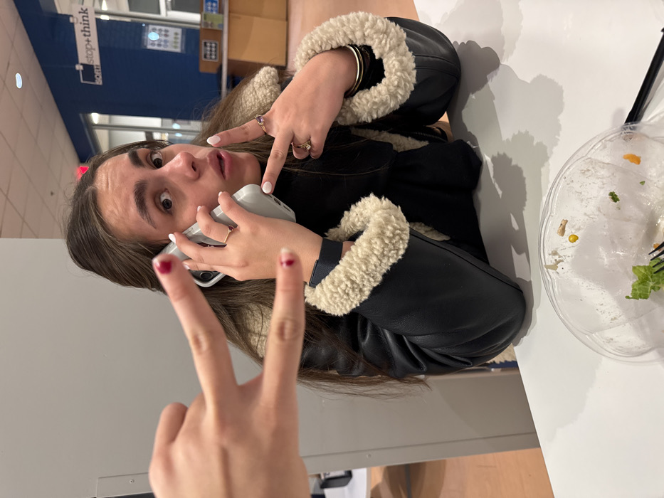 A young woman is seated at a table, wearing a black jacket with a white fur collar. She is talking on a white cell phone and has one hand gesturing with two fingers held up in a peace sign. She has dark hair and is looking at the camera. An empty plastic salad container and a straw are on the table next to her. Behind her, there are blue walls and a sign that says "stop+think".